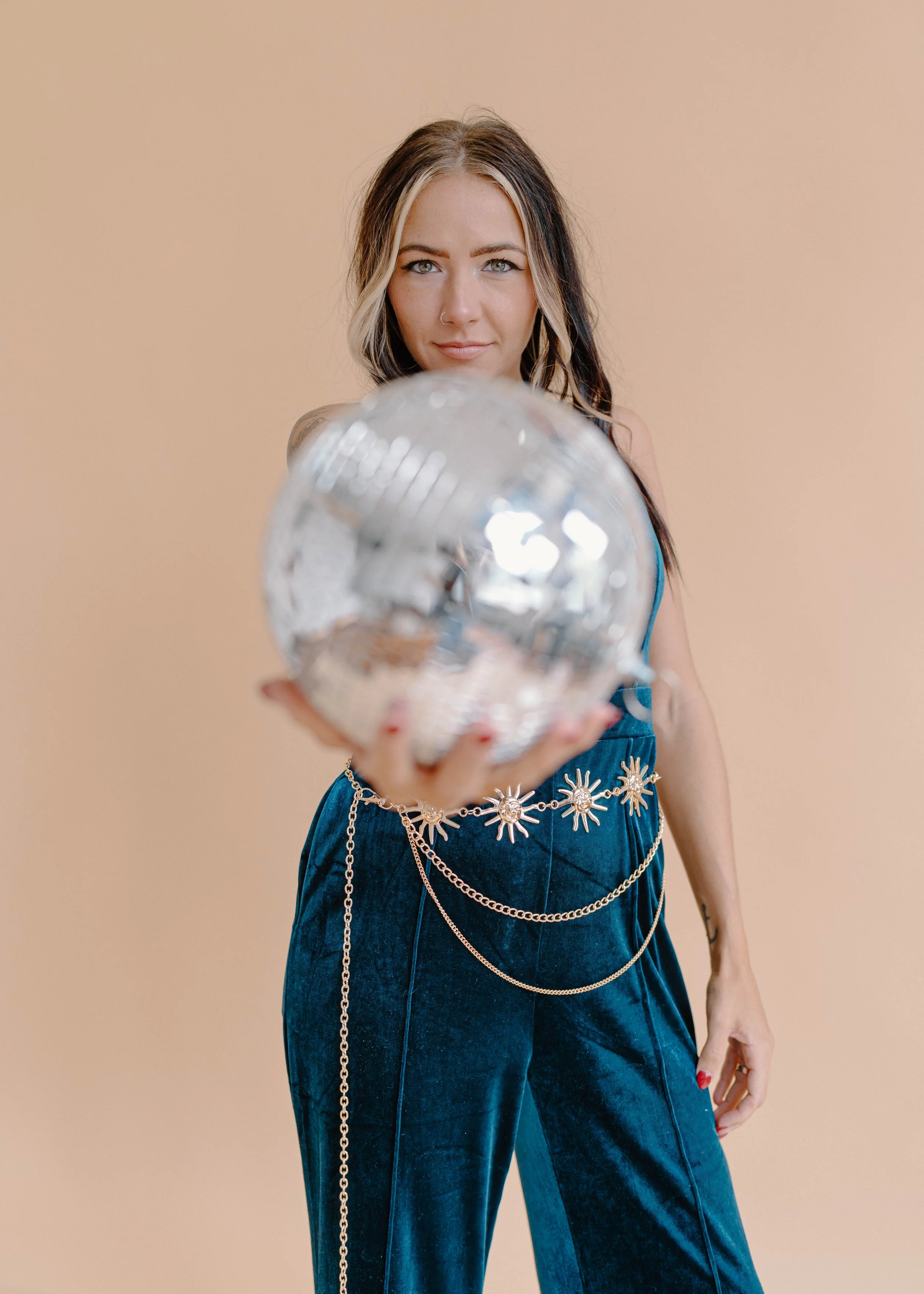 A woman with black and blonde hair holds a clear glass object towards the camera, wearing a blue dress with metallic chain accessories and a beige background.