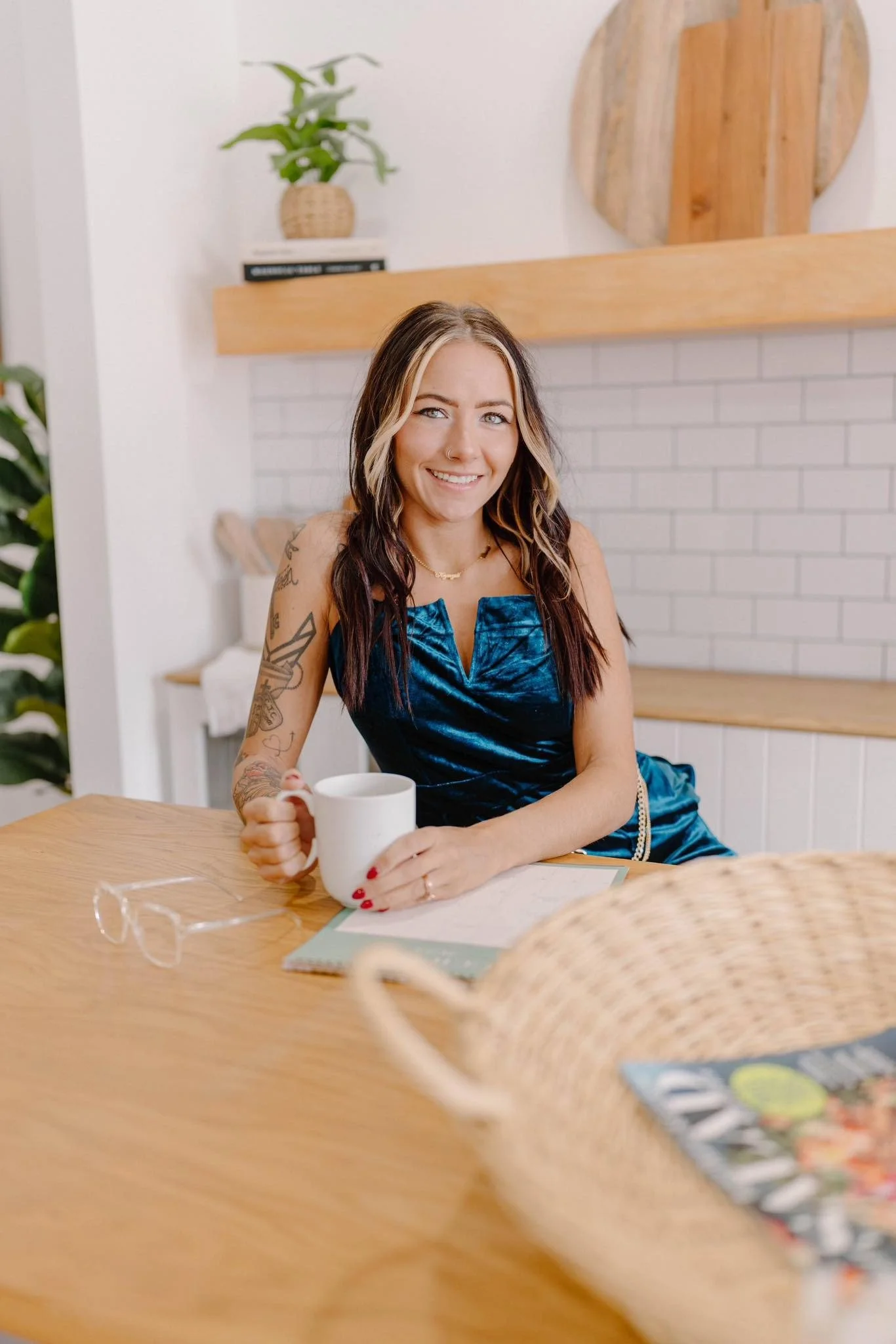 A woman with tattoos on her arm, sitting at a wooden table, smiling, holding a white mug, wearing a blue dress, in a cozy bright room with white brick wall, wooden shelf, and green plants.