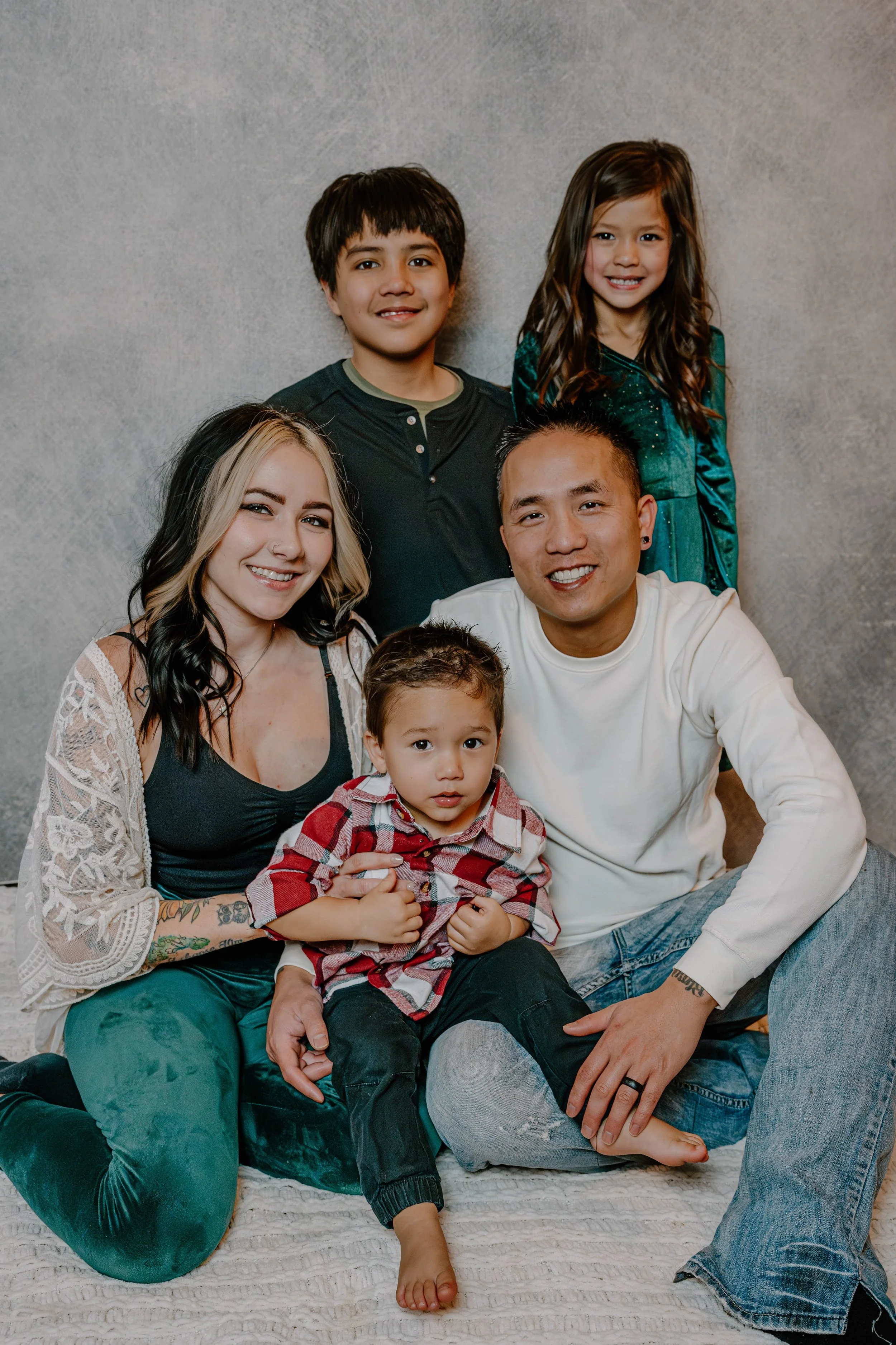 A diverse family of six, including two young children, posing together in a casual indoor setting against a gray textured background. They are smiling and sitting closely, with the adults behind and the children in front.