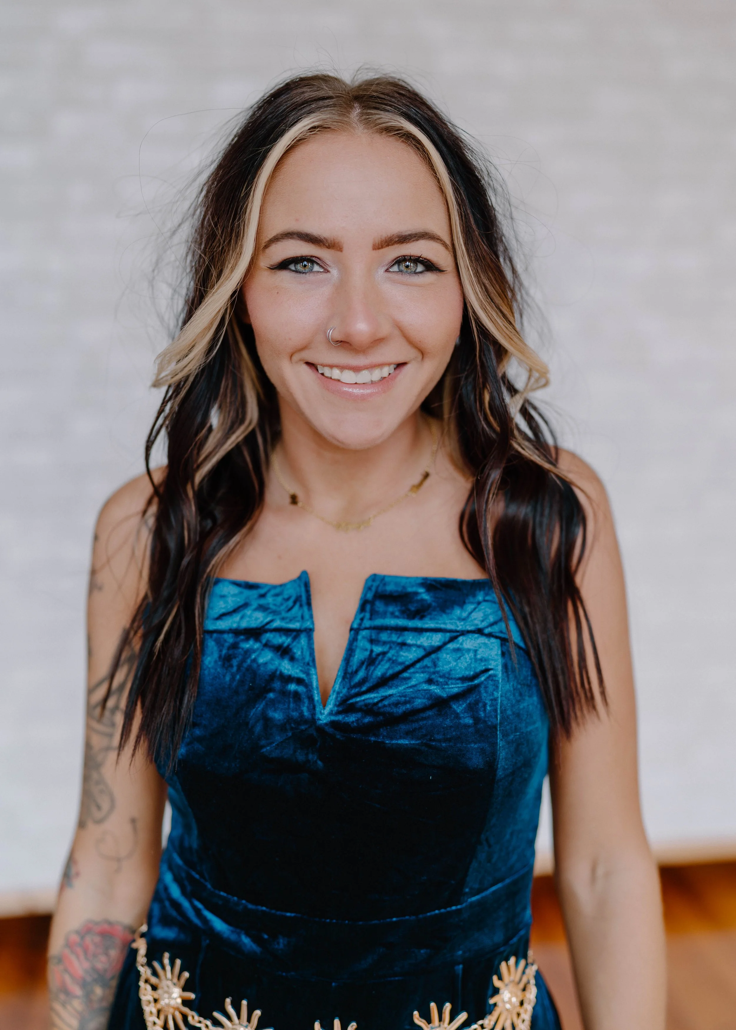 Close-up of a smiling woman with long dark hair and blonde highlights, wearing a blue velvet top, jewelry, and visible tattoos on her right arm.