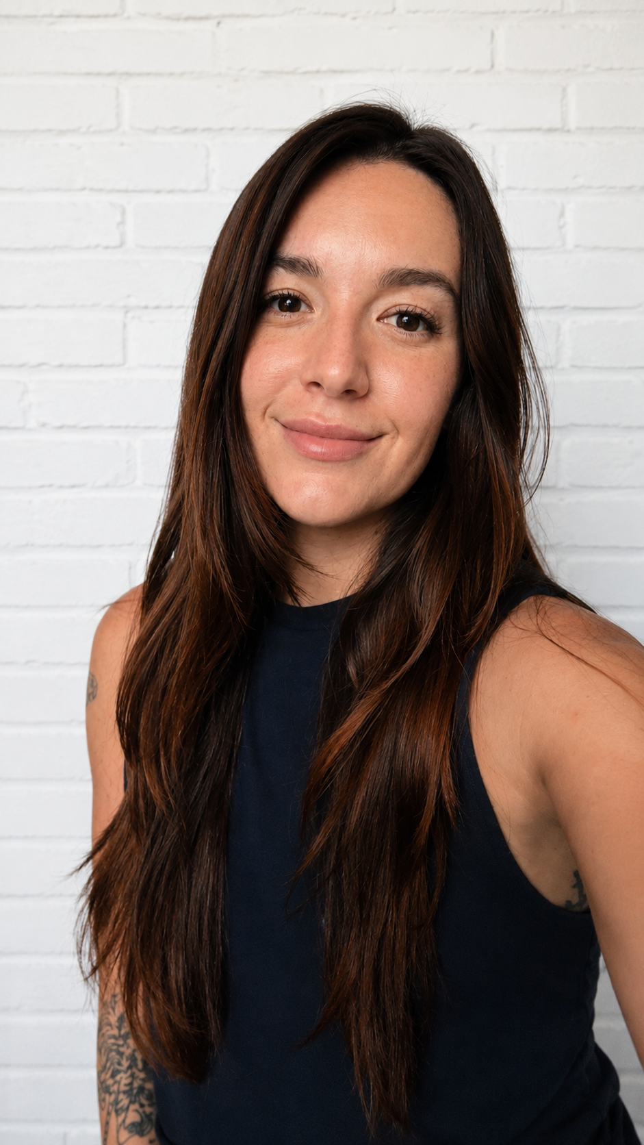 A young woman with long brown hair smiling in front of a white brick wall, wearing a black sleeveless top.