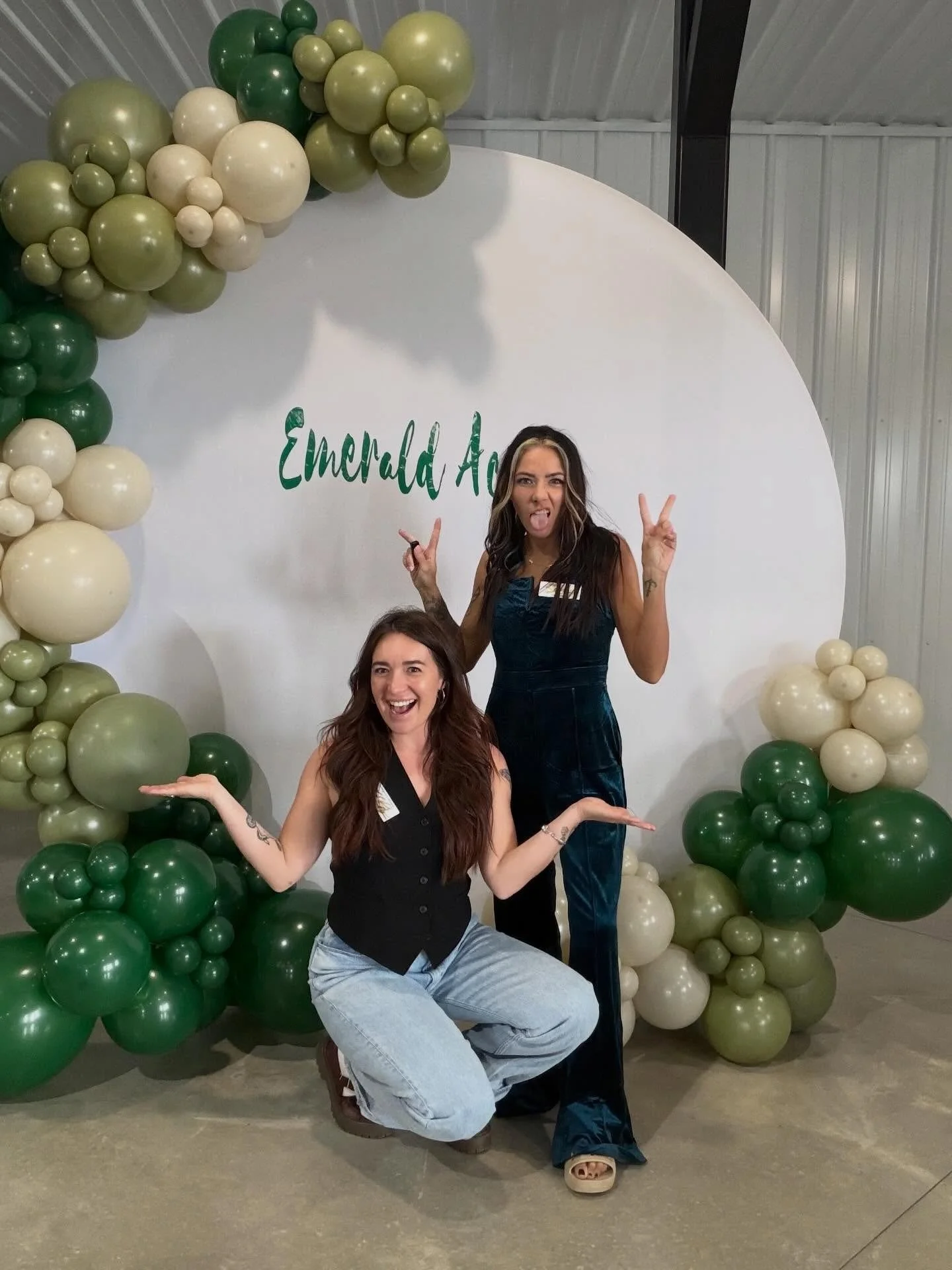 Two women posing in front of a decorative backdrop with green, cream, and beige balloons and the text 'Emerald' partially visible. One woman is squatting with arms out to the sides, smiling, while the other is standing making a peace sign and sticking her tongue out.