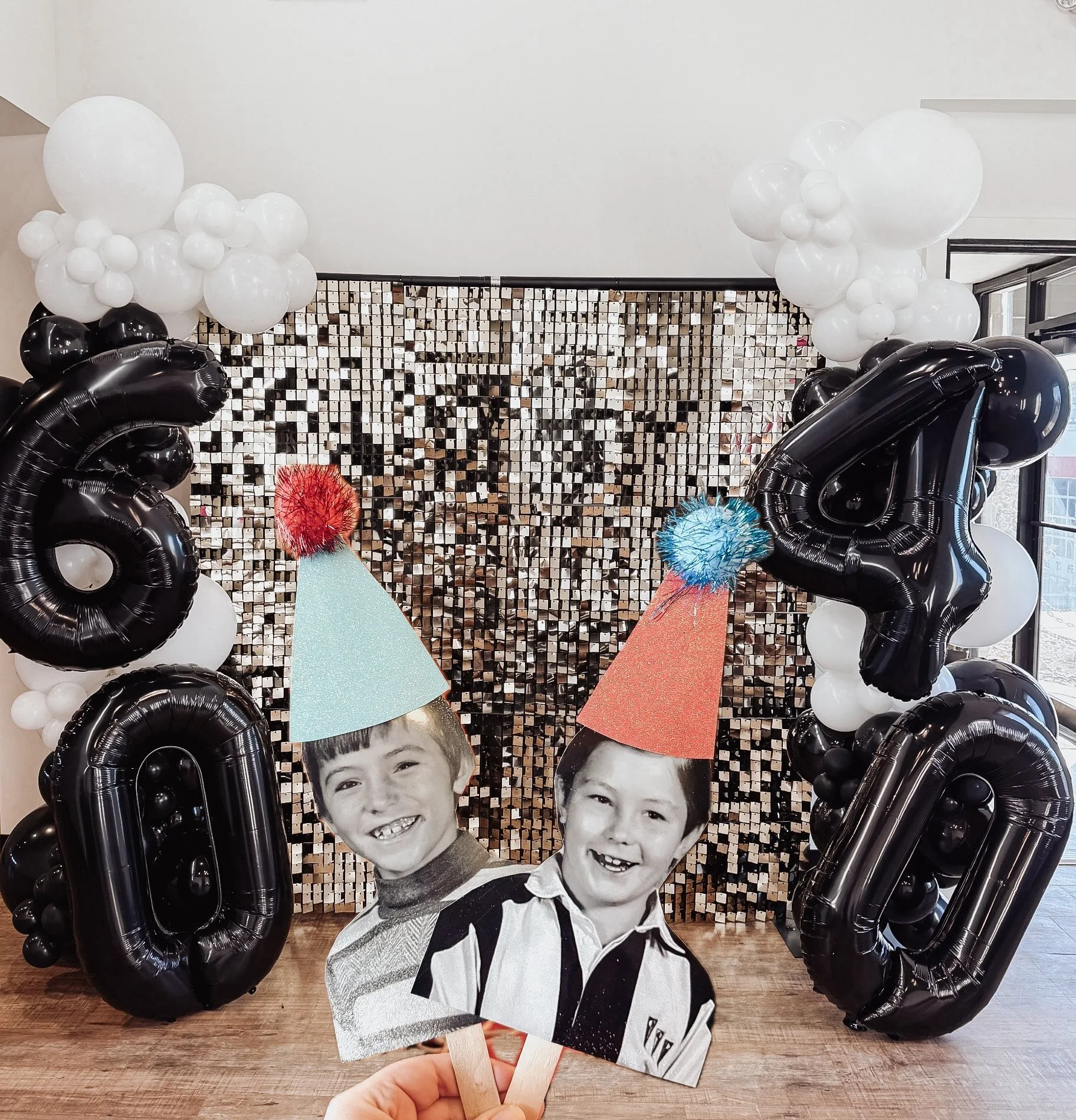 A decorated photo backdrop with black and white balloons and large black number balloons. Hand holding black-and-white photos of smiling children in front of the backdrop.