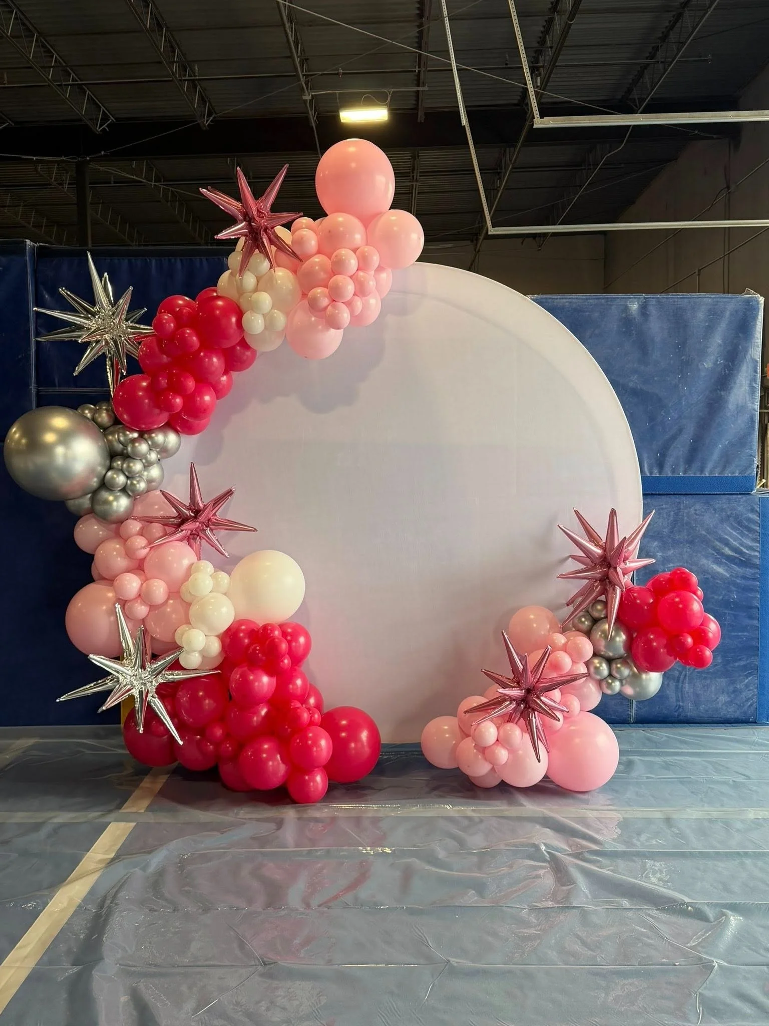 Decorative balloon arch with pink, red, white, and silver balloons and star-shaped balloons, set against a backdrop with a white circle on a blue background in an indoor space.