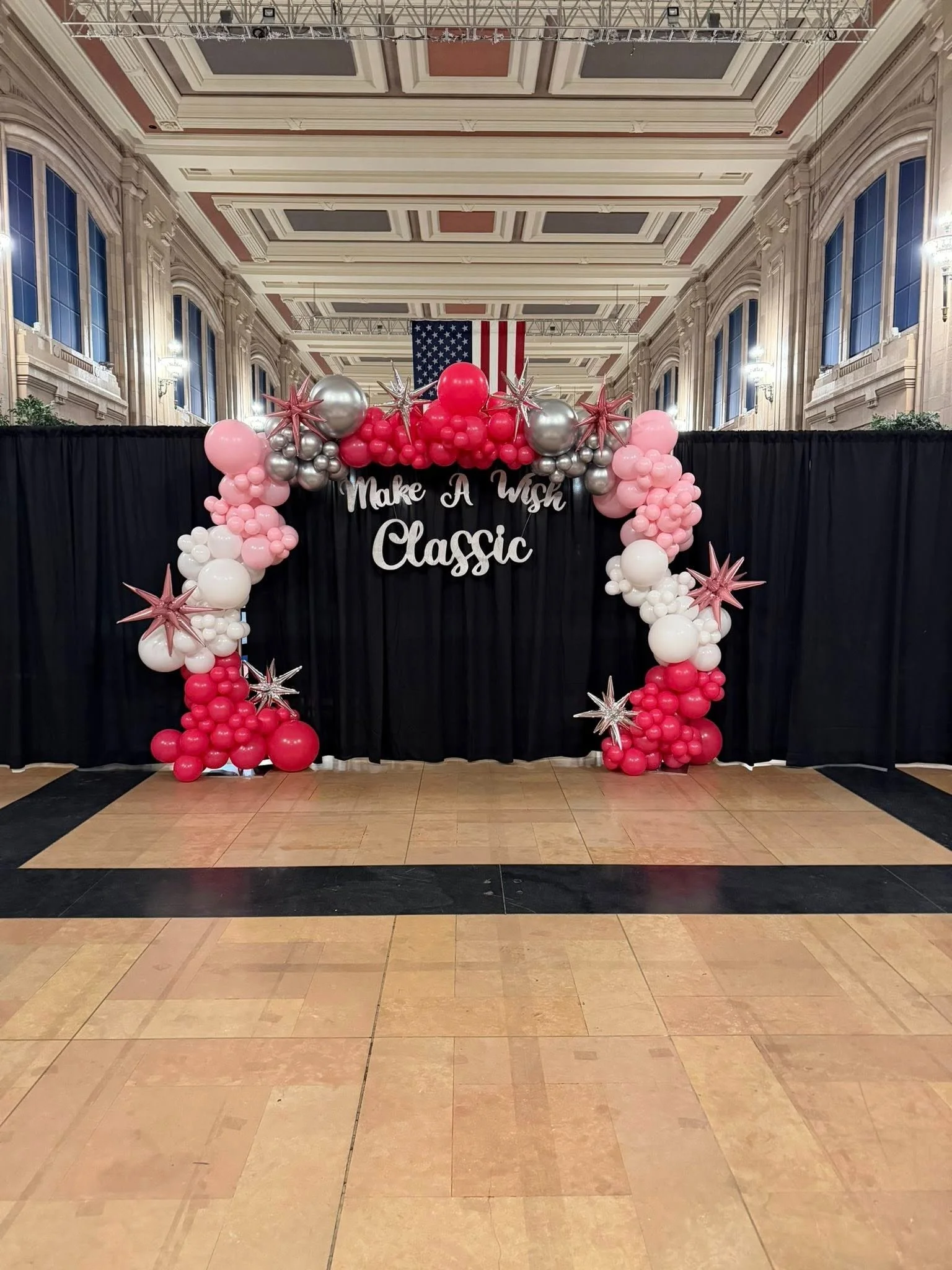 Balloon arch with pink, white, red, and silver balloons, featuring star-shaped balloons, in front of a black curtain with a sign that says "Make A Wish Classic," inside a grand hall with high ceilings, large windows, and an American flag.