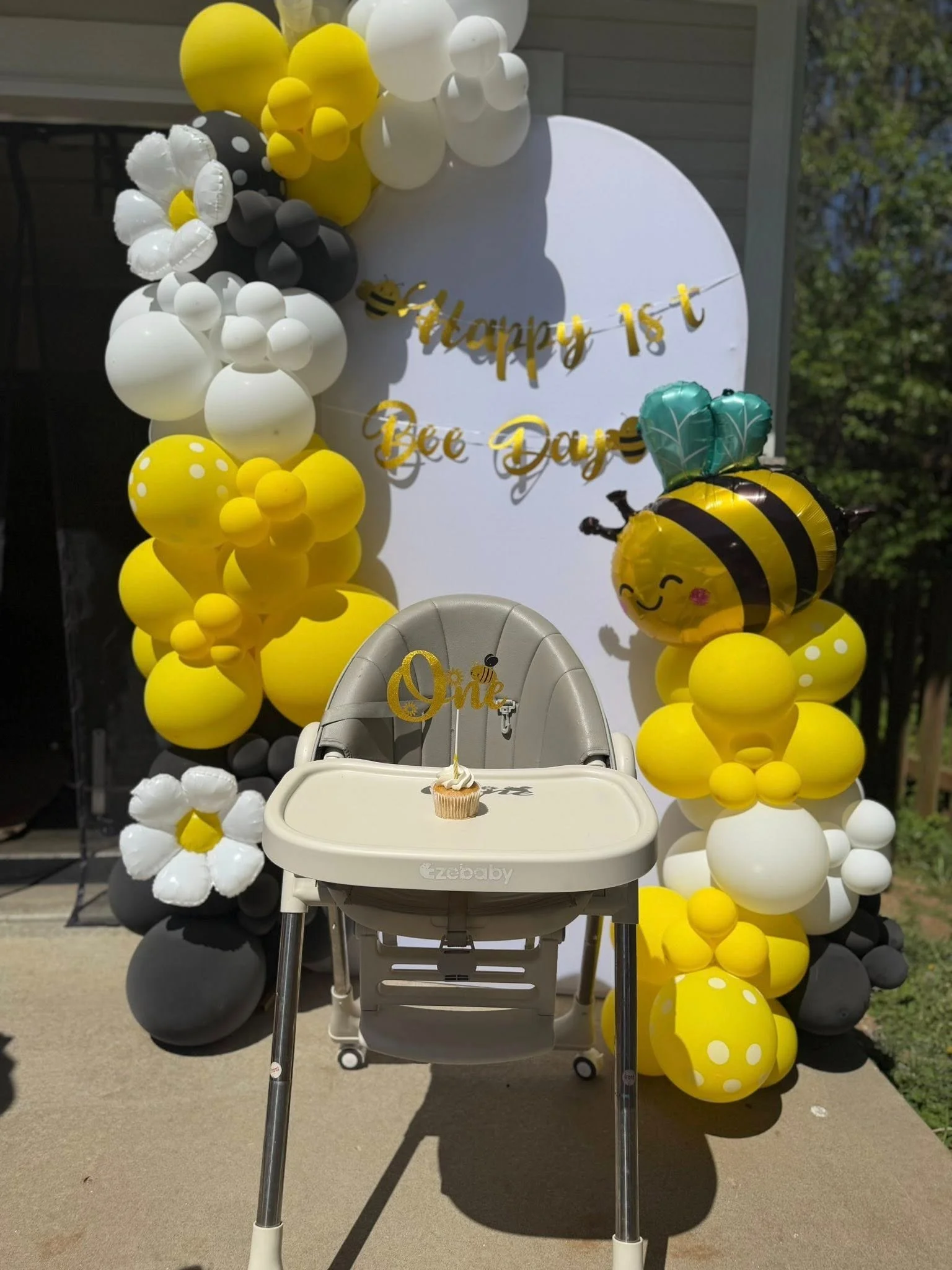 Party decoration with yellow, white, and black balloons arranged around a sign that reads 'Happy 1st Bee Day' and a cute bee-shaped balloon. A cupcake with a single candle is in front of a white high chair.
