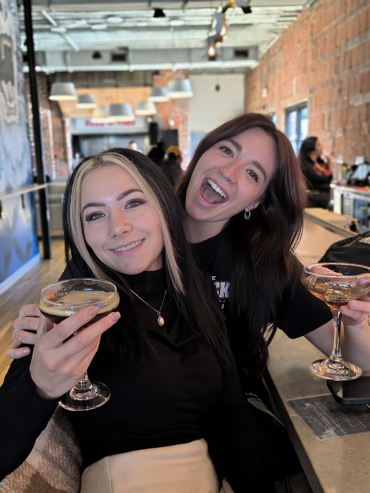 Two women smiling and celebrating with cocktails at a restaurant or bar with exposed brick walls, pendant lighting, and a modern industrial interior.