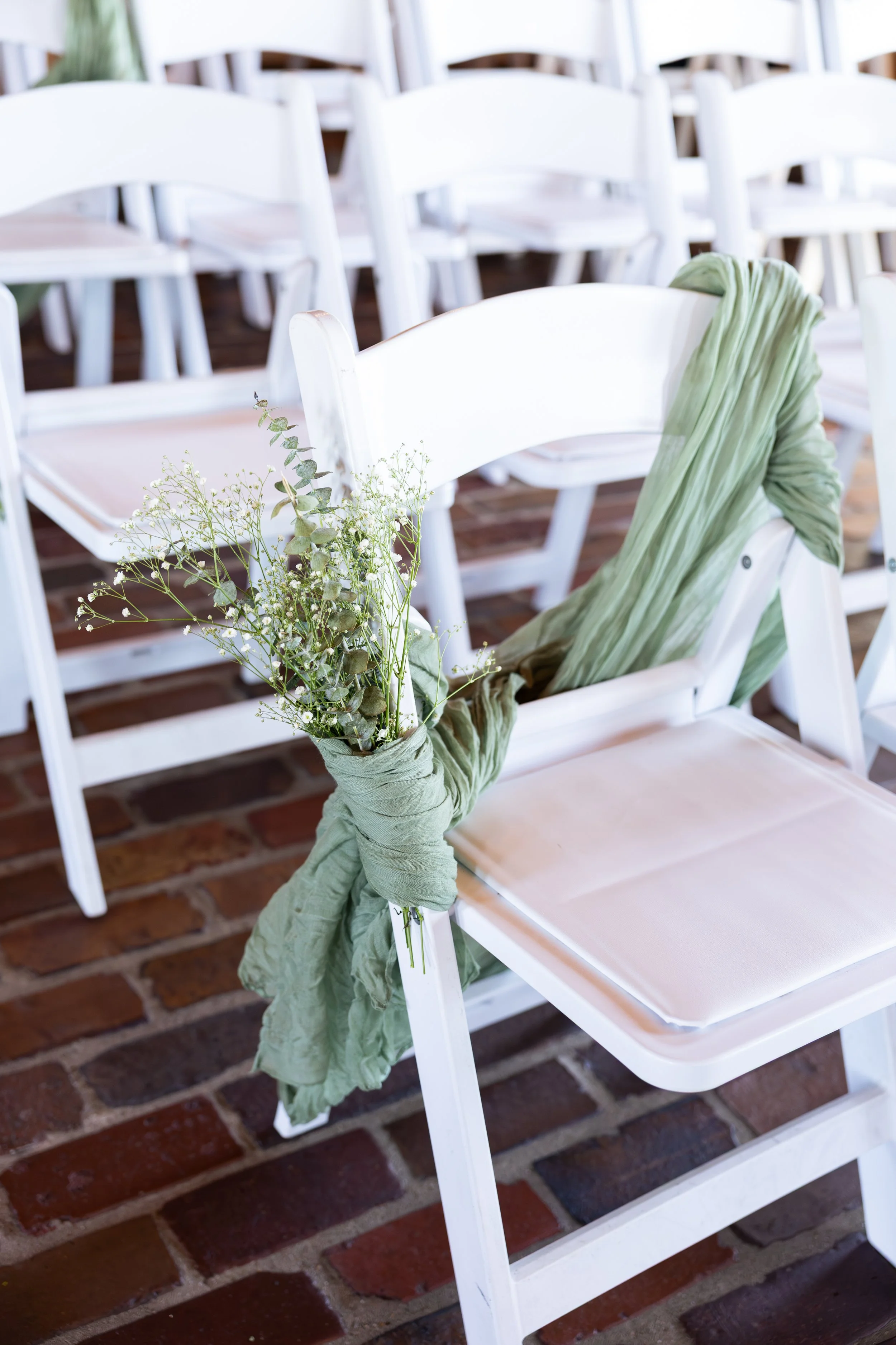 White chairs decorated for a wedding, with green fabric and a small bouquet of white and green flowers tied to the back of each chair.