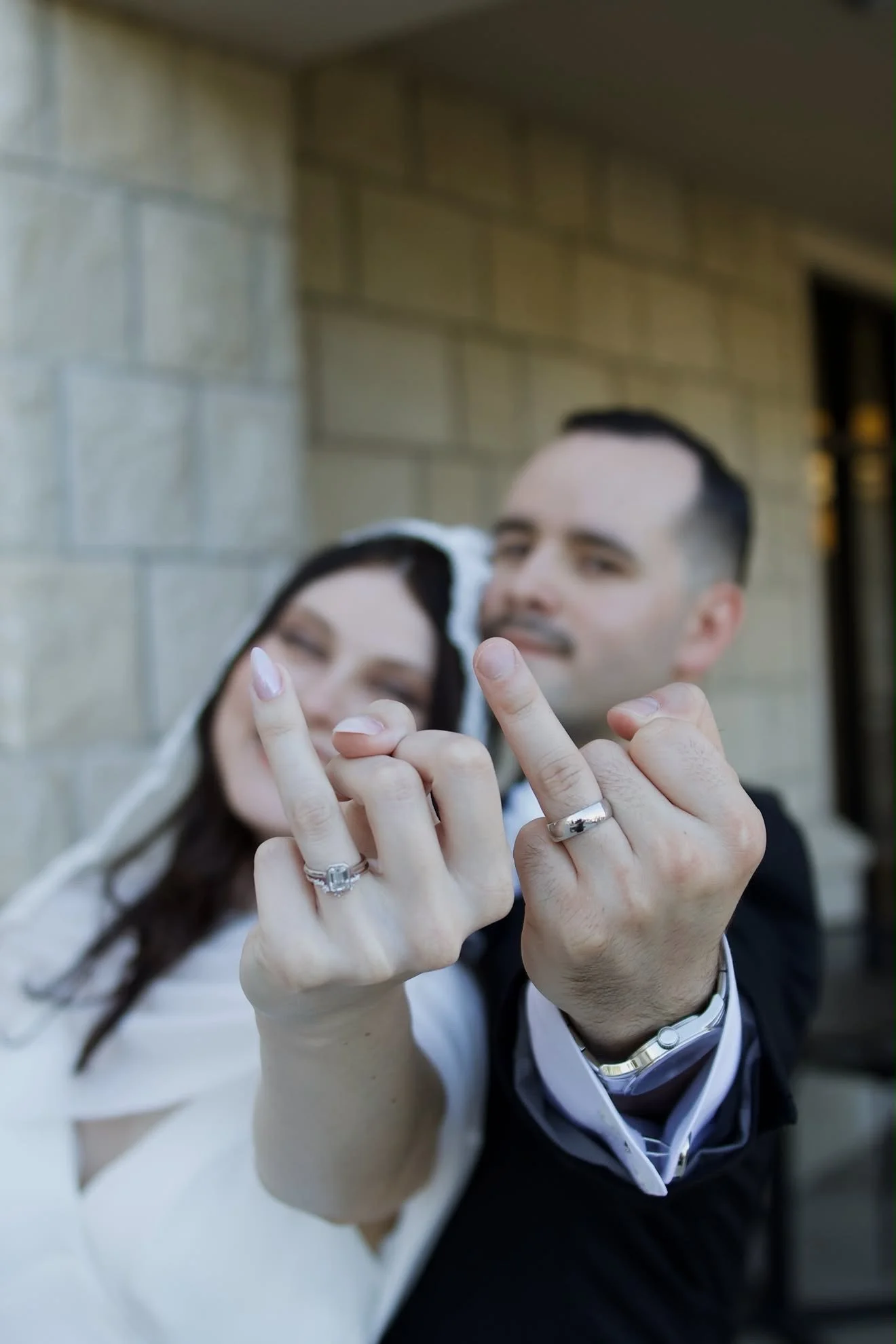 A couple showing their wedding rings while taking a selfie outdoors, with a brick wall in the background.