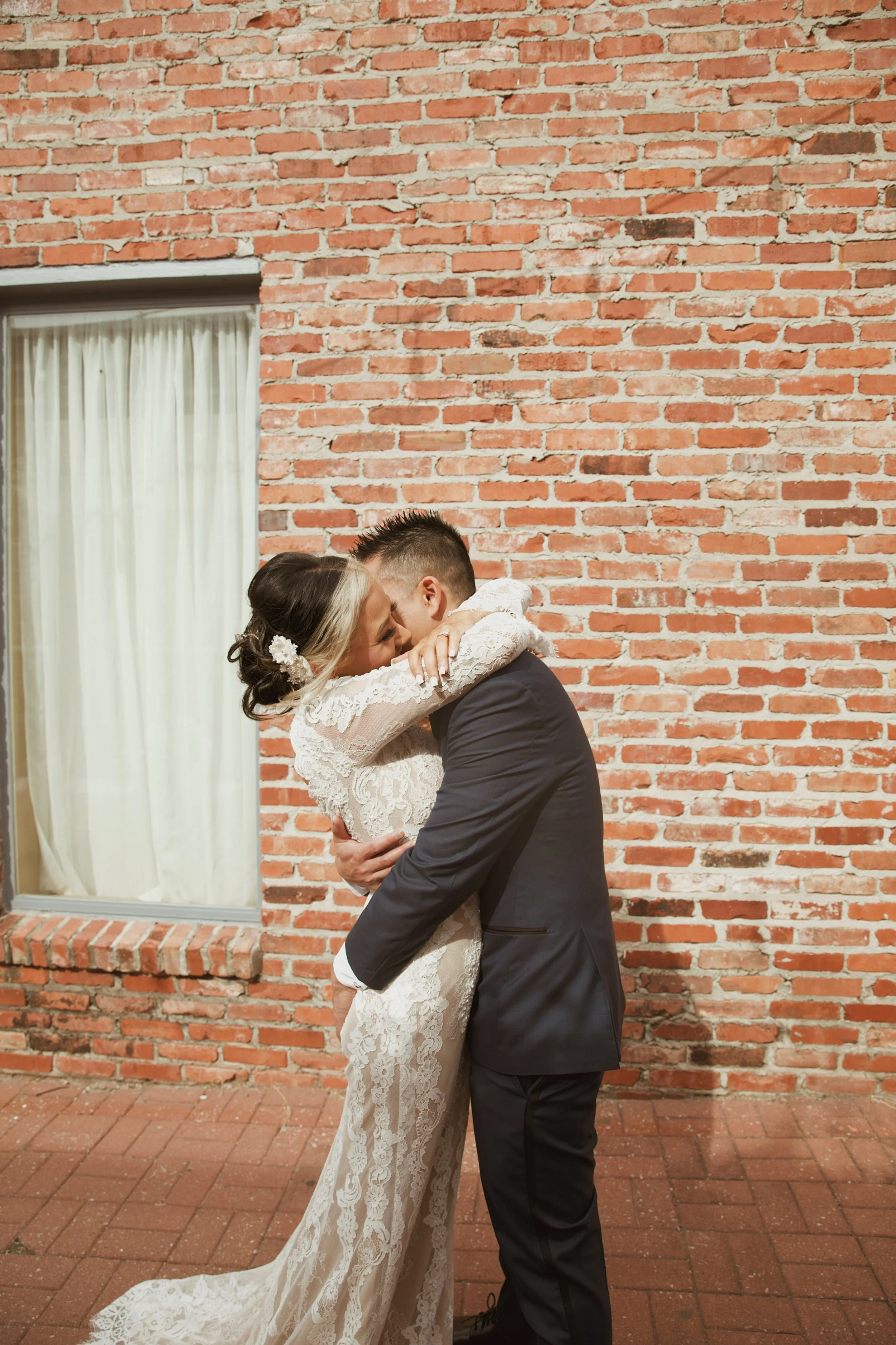 A newlywed couple embraces and kisses in front of a brick wall with a window that has white curtains.