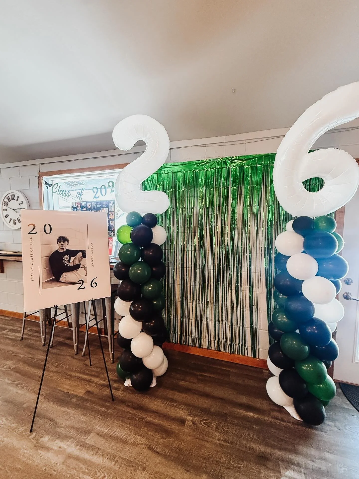 Decorations for a graduation celebration with large white number balloons forming '20', flower balloon columns in green, black, white, and blue, a backdrop with green metallic fringe, and a poster with a photo of a young man, the text 'Class of 2020' and the date 'May 26'.