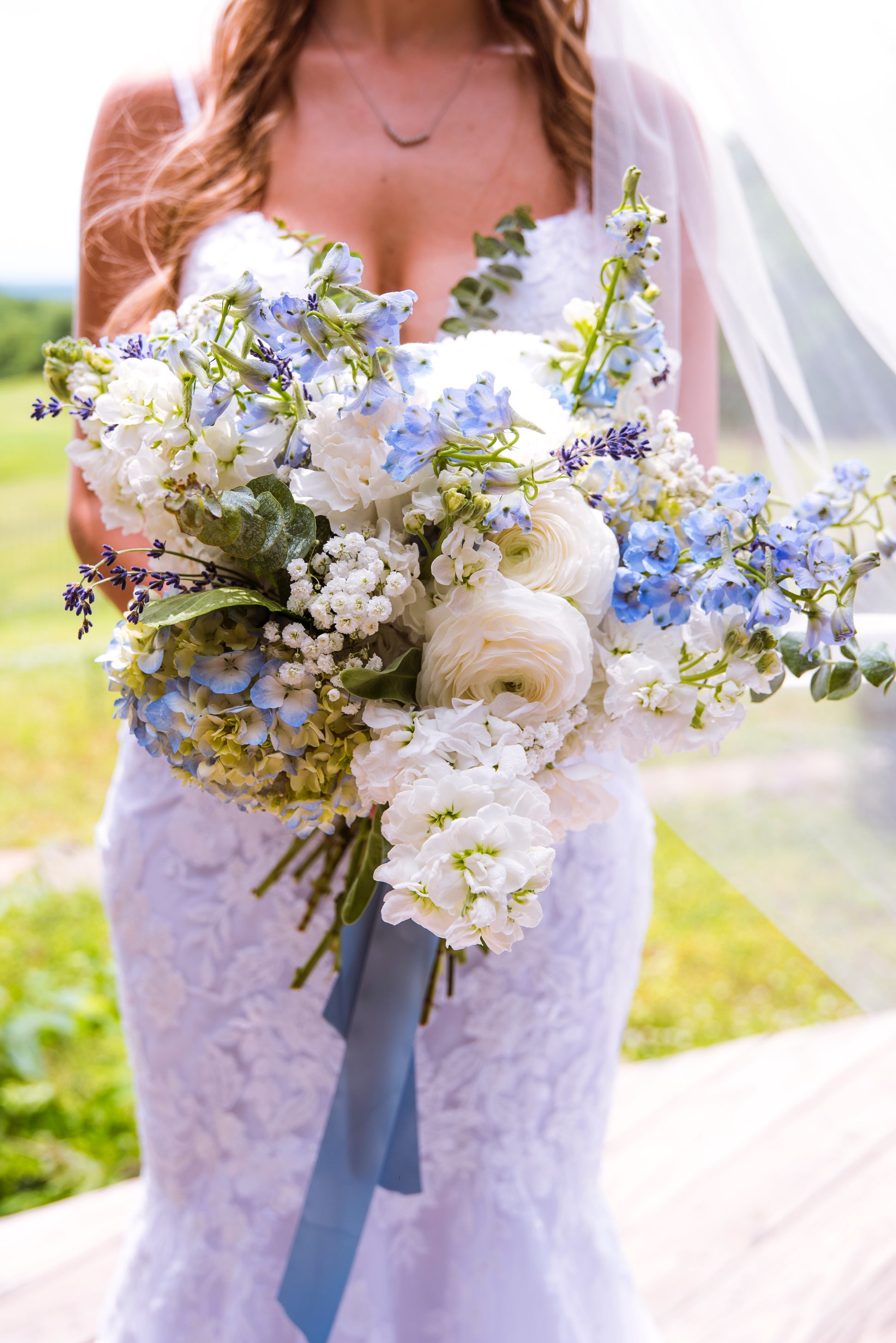 A woman in a white lace dress holding a large bouquet of white and blue flowers, outdoors on a sunny day.