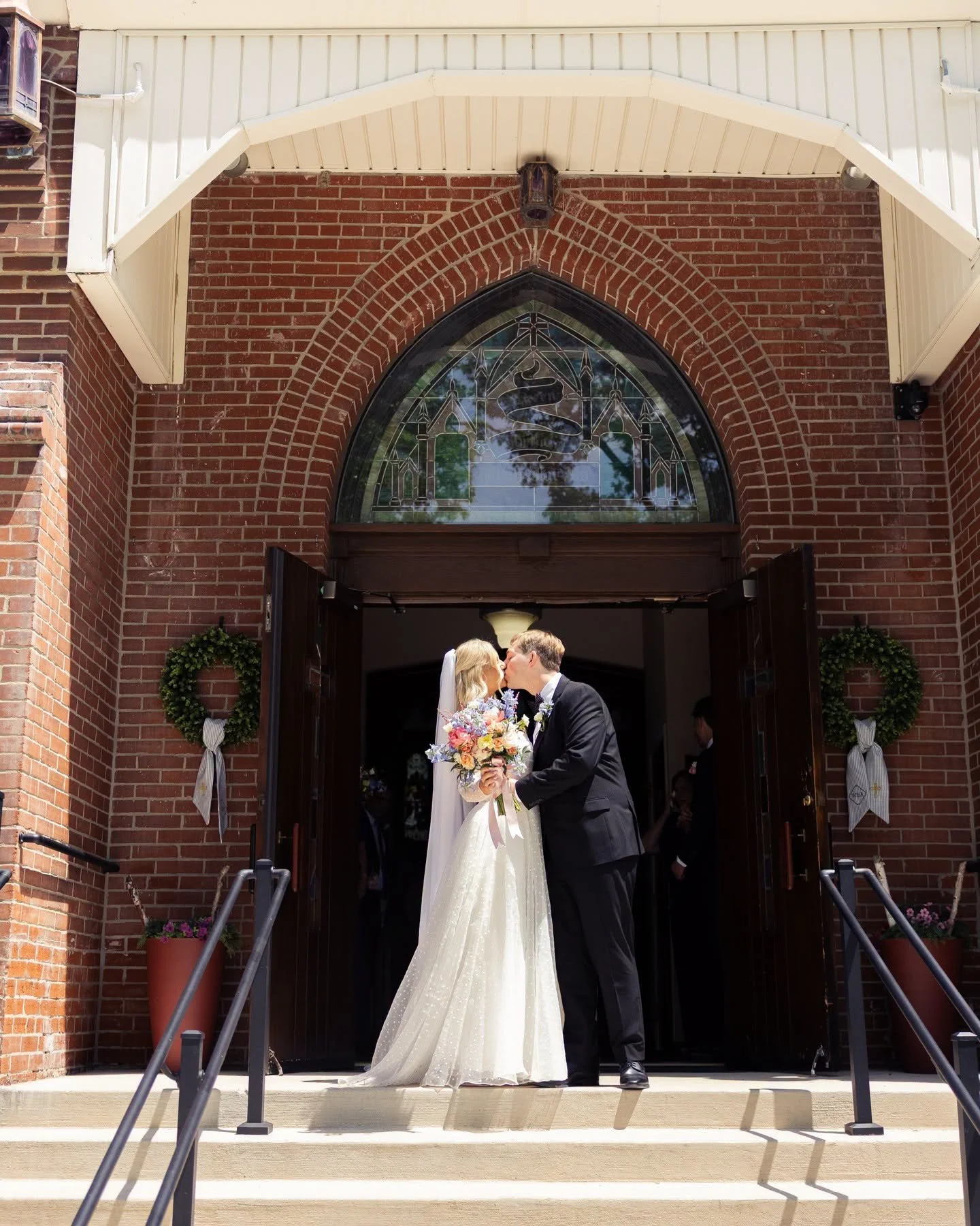 Bride and groom sharing a kiss outside a church after their wedding, holding a bouquet of flowers, with church doors open behind them, floral wreaths on either side of the entrance, and people inside the church.