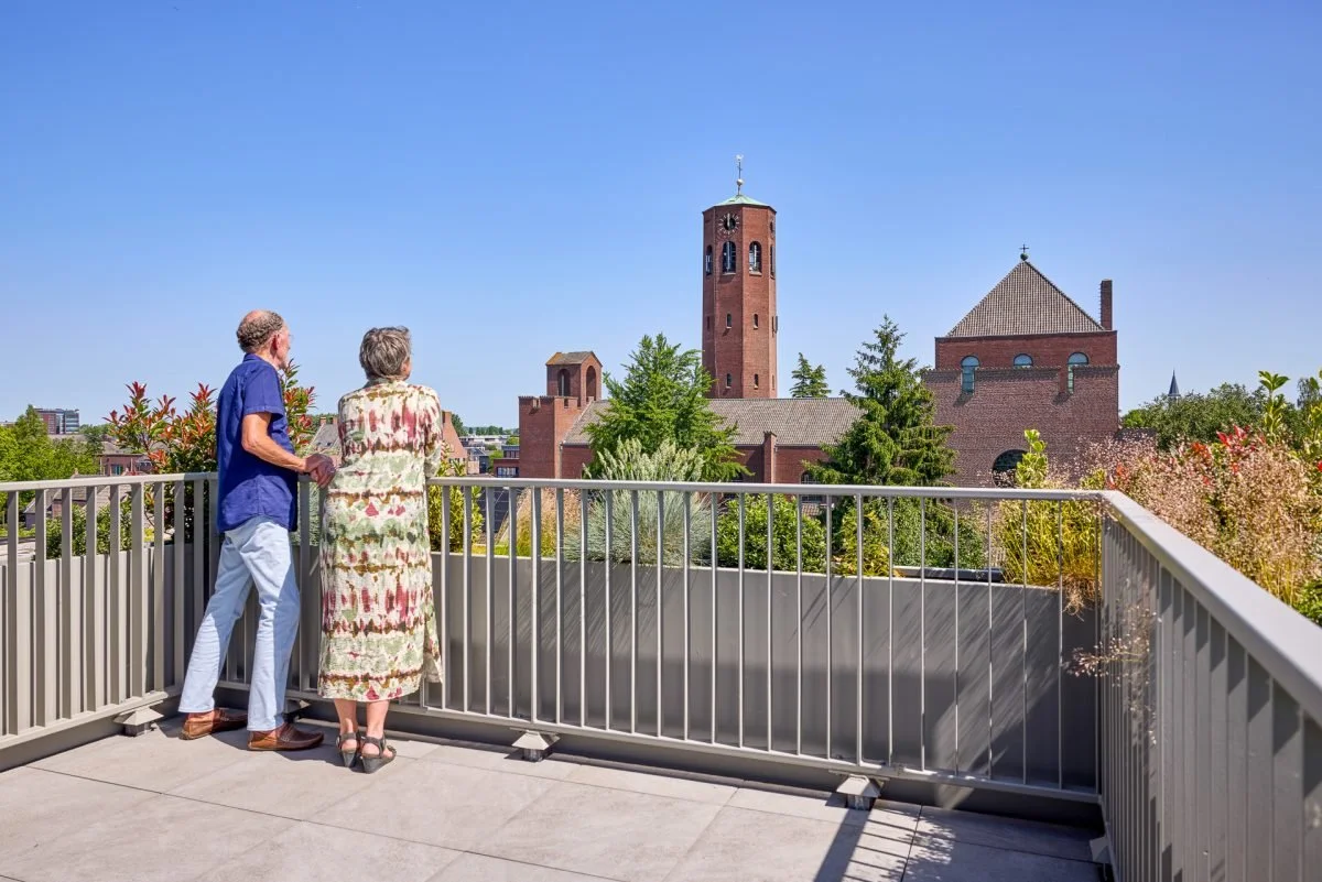 Twee oudere mensen kijken vanaf een balkon naar een kerktoren en een kerk in een stad met veel groen onder een blauwe lucht.