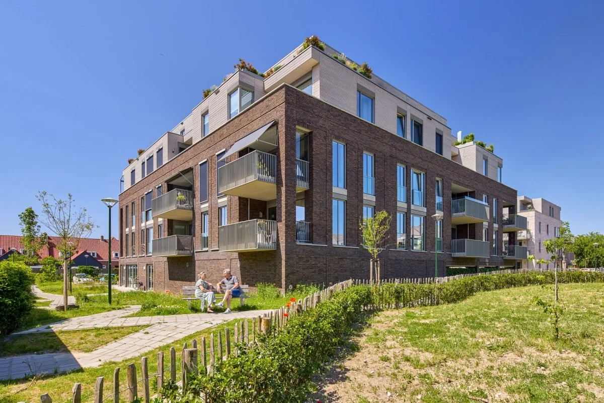 Twee mensen zitten op een bankje in een groen park voor een modern, meerlaagse appartementencomplex met grote ramen en balkons, onder een heldere blauwe hemel.