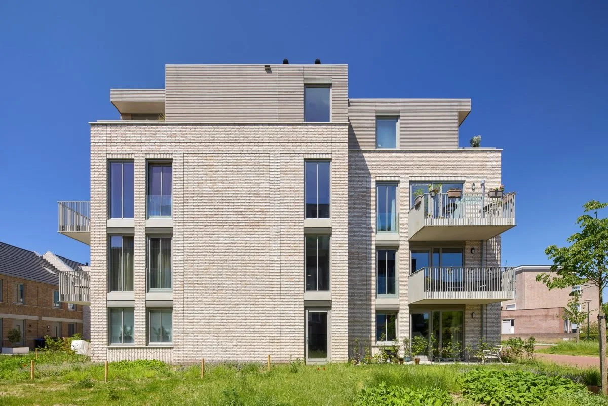 Moderne, vier verdiepingen tellende appartementencomplex met grote ramen, balkons en een lichte bakstenen gevel, omgeven door een grasveld en blauwe lucht.