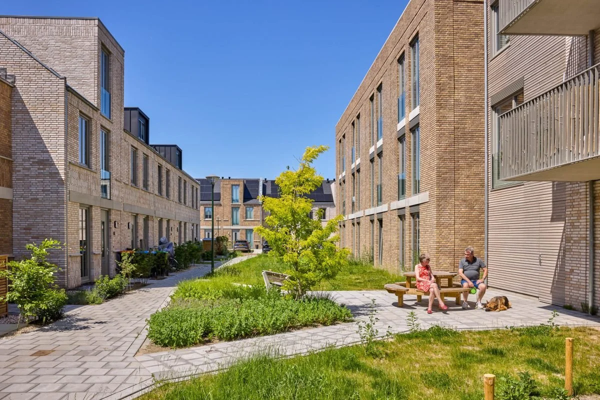 Twee mensen zitten op een bank in een binnenplaats tussen moderne appartementen. Een hond ligt op de grond naast ze. Er is een boom en groen gras, en helderblauwe lucht.