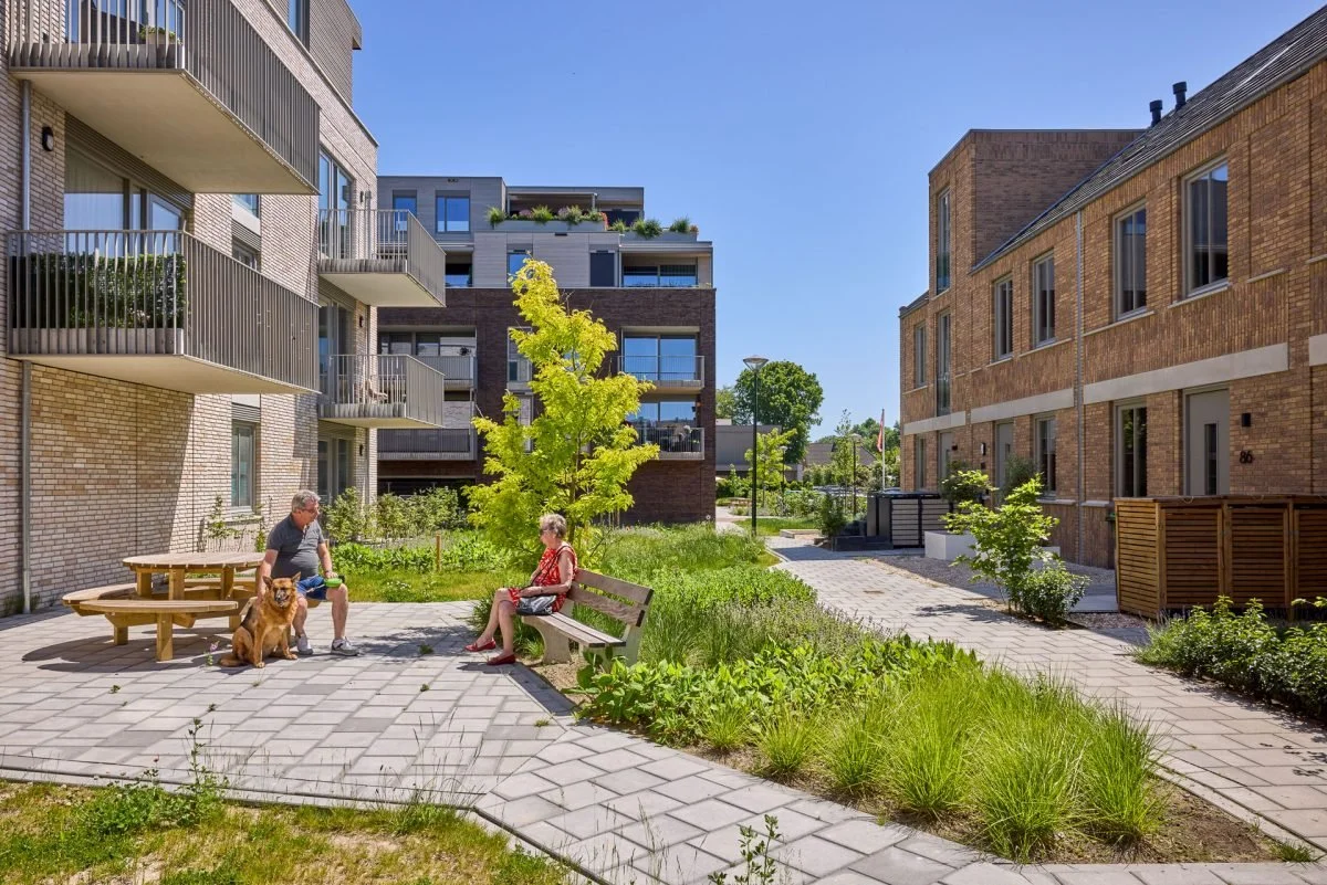 Twee mensen zitten in een groene binnenplaats met bankjes en planten, een man met een hond en een vrouw die op een bankje zit op een zonnige dag, omringd door moderne appartementen.