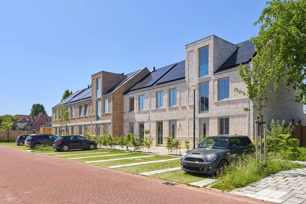 Moderne rijtjeshuizen met zonnepanelen op het dak, geparkeerde auto's voor de huizen, groene bomen en een blauwe lucht.