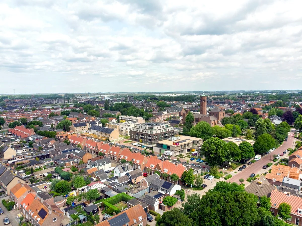 Ingezonden foto van een stadsbeeld met een mix van huizen, gebouwen en een kerktoren, omgeven door groene bomen en een weg met geparkeerde auto's onder een bewolkte lucht.