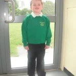 Young boy wearing a green hoodie and black pants standing in front of a glass door