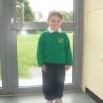 Child in green sweater and dark pants standing near glass door with grass outside.