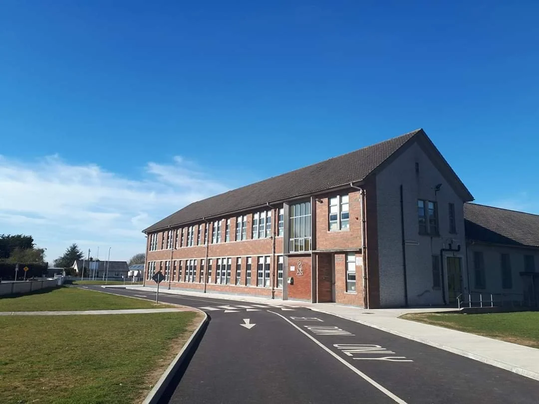 A two-story brick school building with numerous windows, situated on a curved road with marked lanes and a grassy area. The sky is clear and blue.