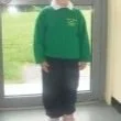 Child in green sweater and dark pants standing near glass door with grass outside.