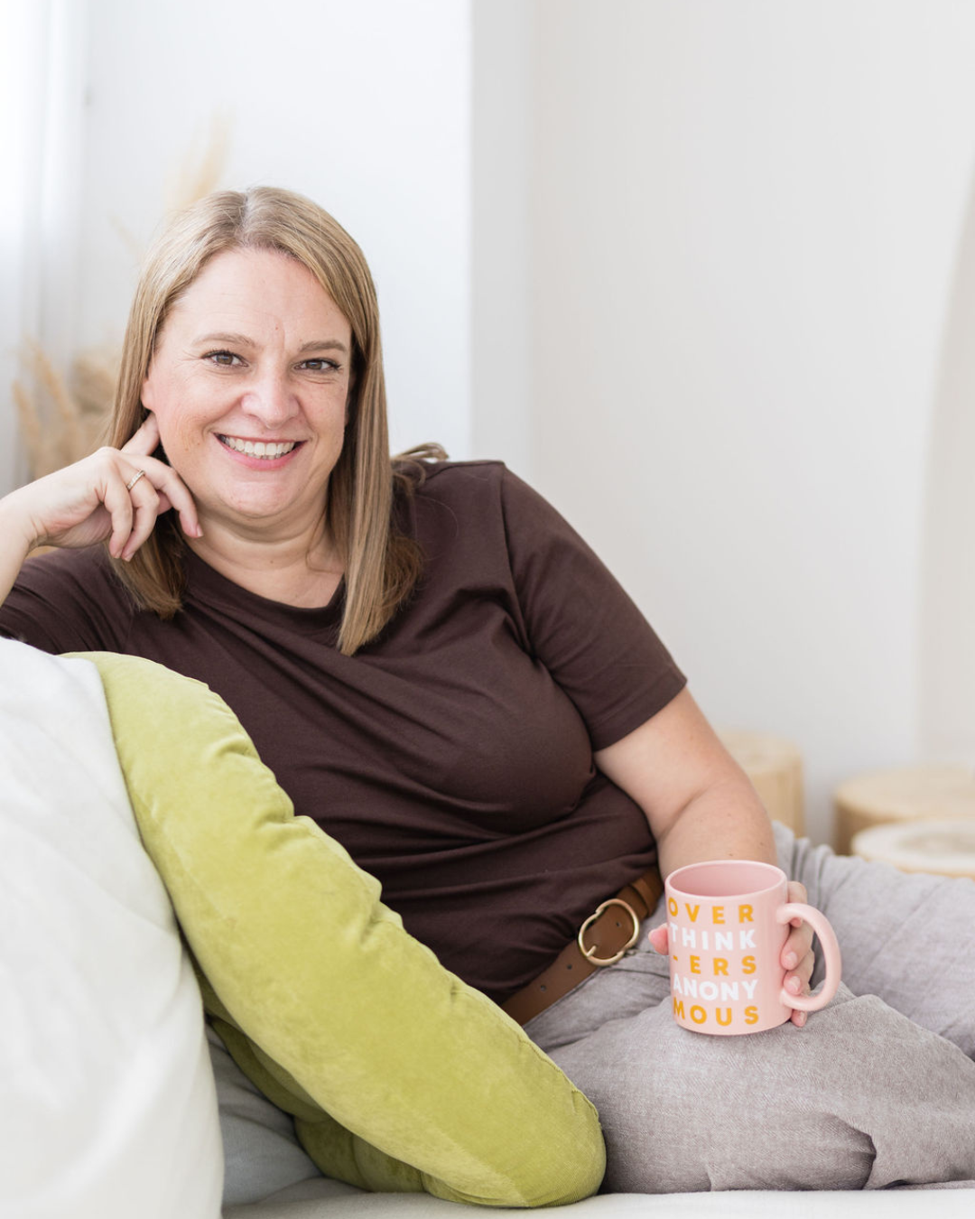 A woman with shoulder-length blond hair smiling, sitting on a sofa holding a pink mug with text, and resting her head on her hand, in a relaxed home setting.