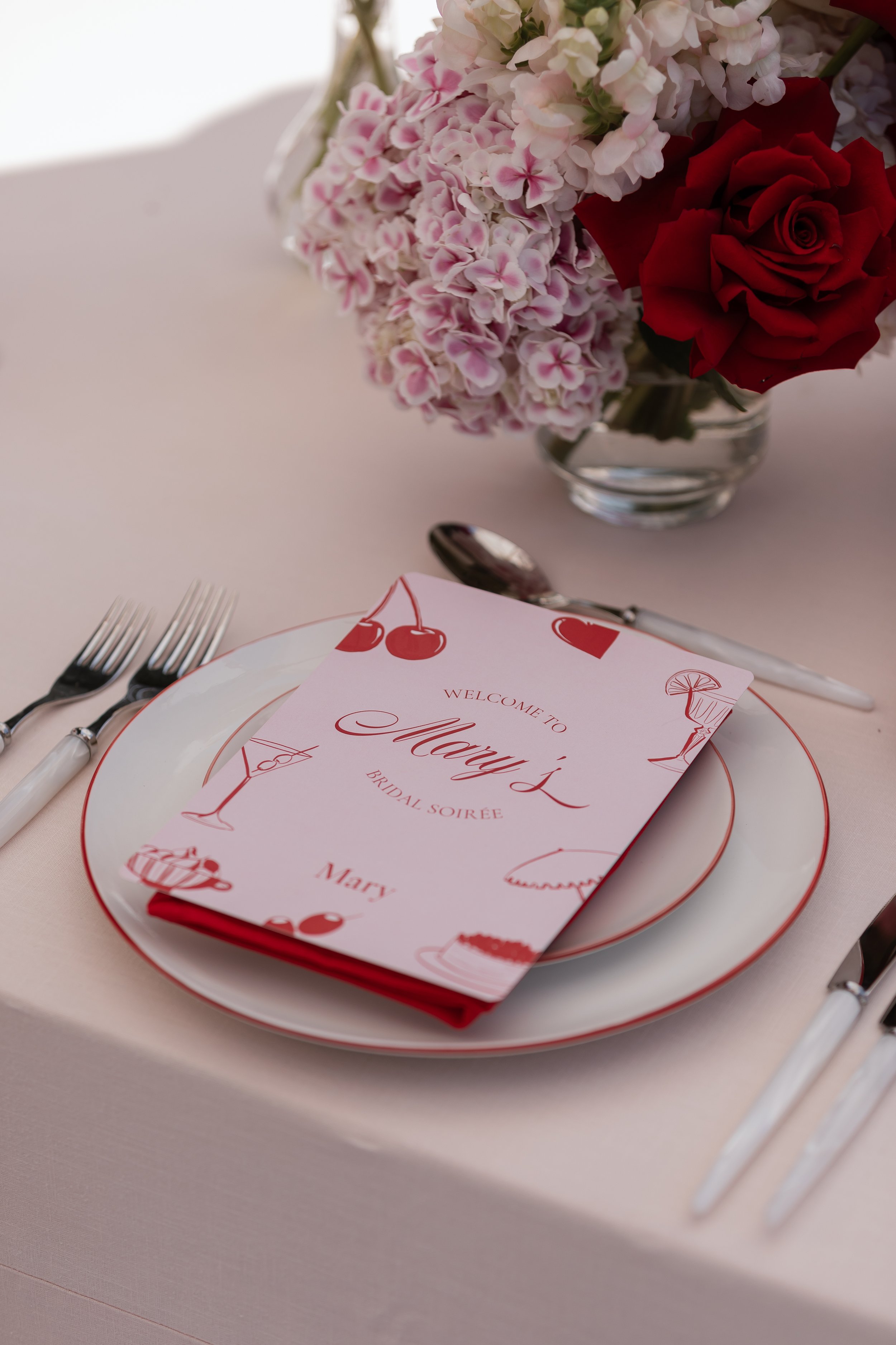 A table setting with a pink and white floral centerpiece and a white plate holding a folded napkin with a pink and white paper menu on top. The menu has illustrations of a cherry, a martini glass, and a slice of lemon, and the word 'Merry' is visible.
