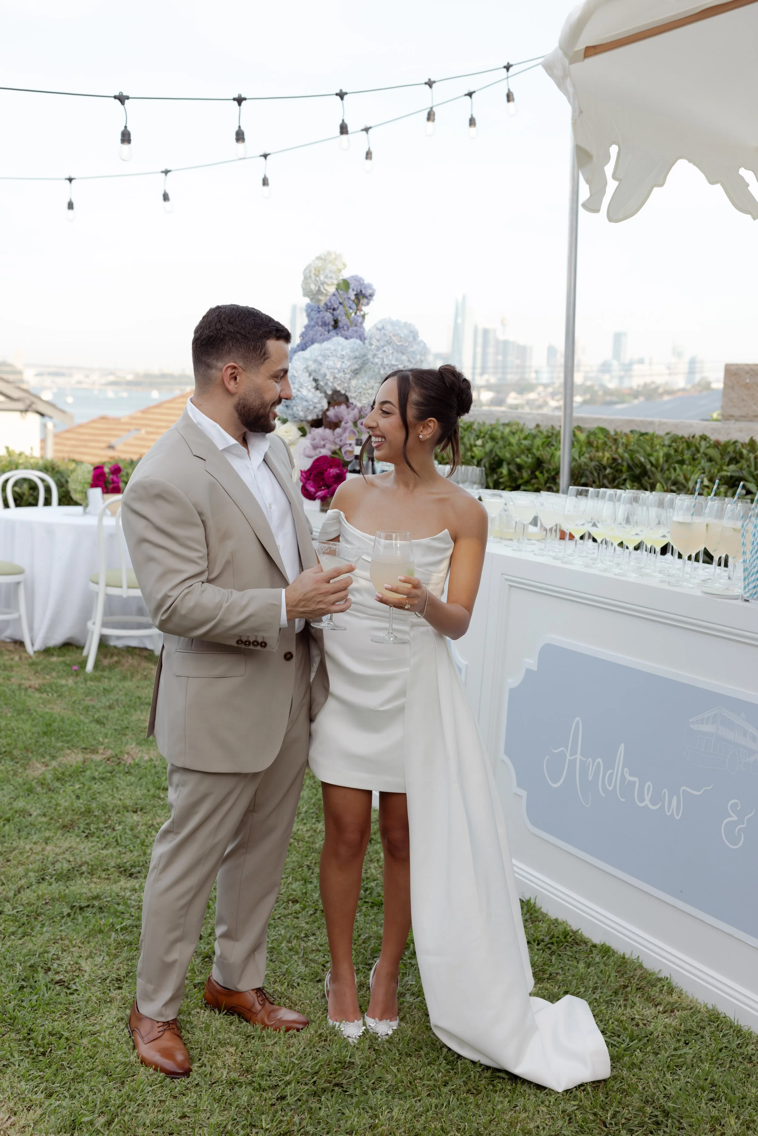 An engaged couple smiling at each other holding glasses of drinks at an outdoor engagement reception under string lights, with cityscape in the background.