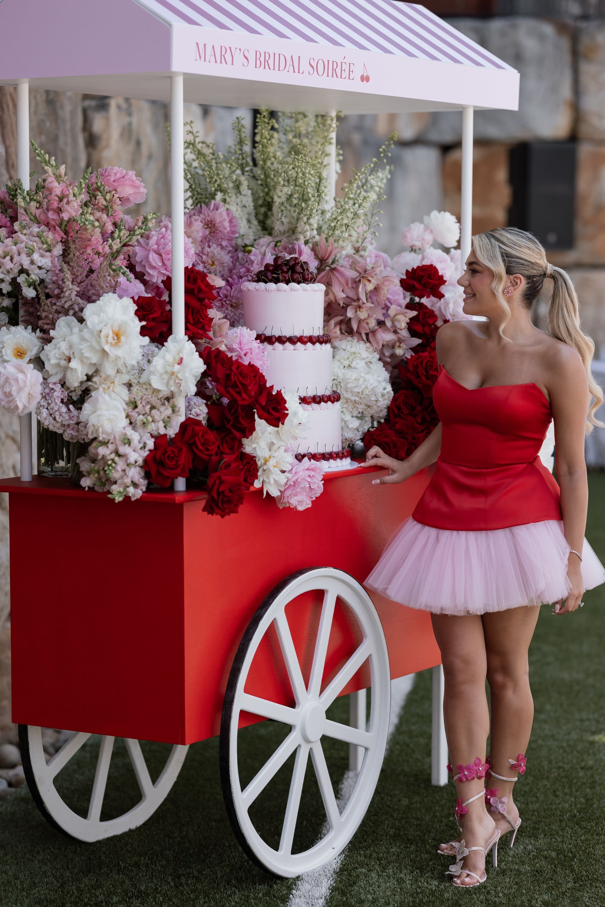A woman in a red strapless top and pink tulle skirt stands next to a floral cart with a sign that reads "Mary's Bridal Soiree". The cart is decorated with pink, red, white, and green flowers and has a three-tiered pink cake with cherries on top.
