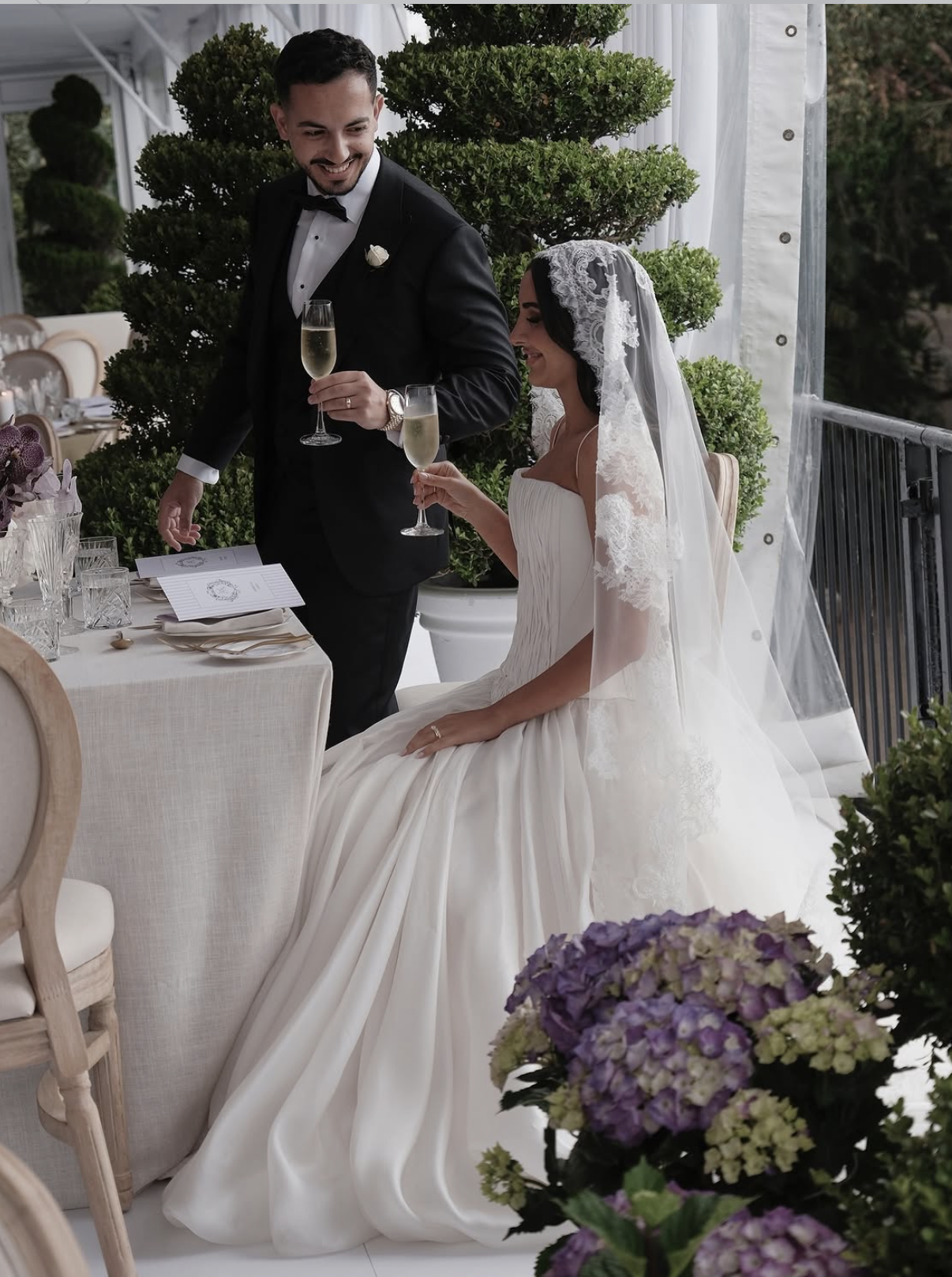 A bride and groom celebrating at their wedding reception, holding champagne glasses. The bride is seated in a white wedding gown with a veil, and the groom is standing in a black tuxedo. They are surrounded by elegant decor and greenery.