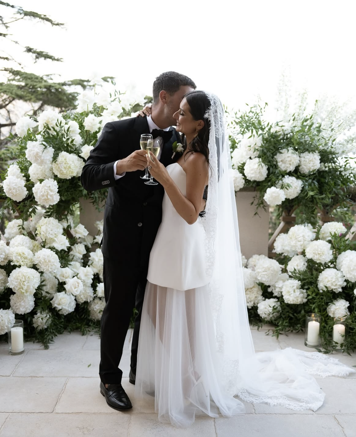 A newlywed couple in wedding attire sharing a kiss and holding champagne glasses in front of a backdrop of white flowers and candles.