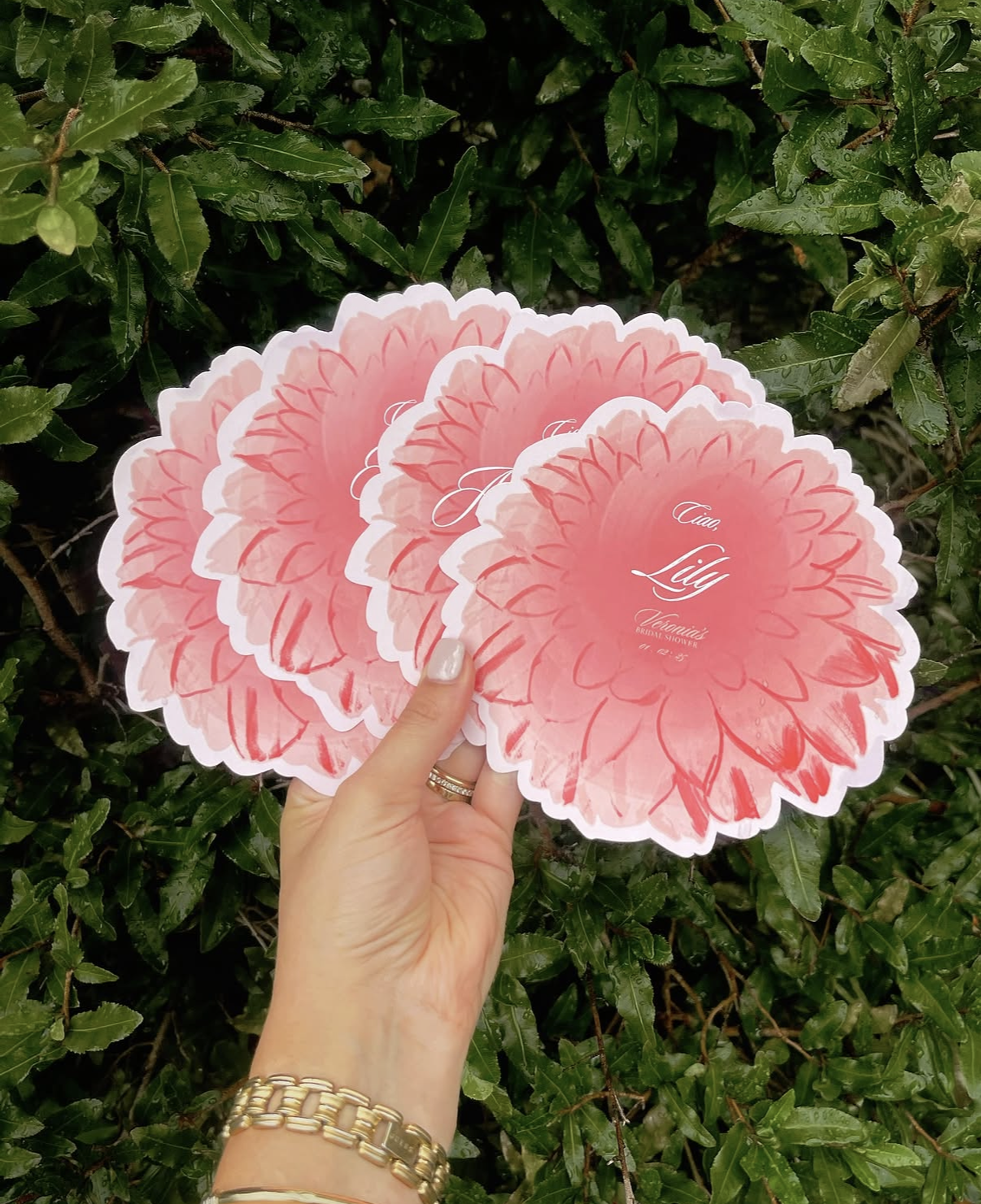 Hand holding three pink flower menu place cards and the words "Ciao, Lily" written on them, in front of green leafy bushes.