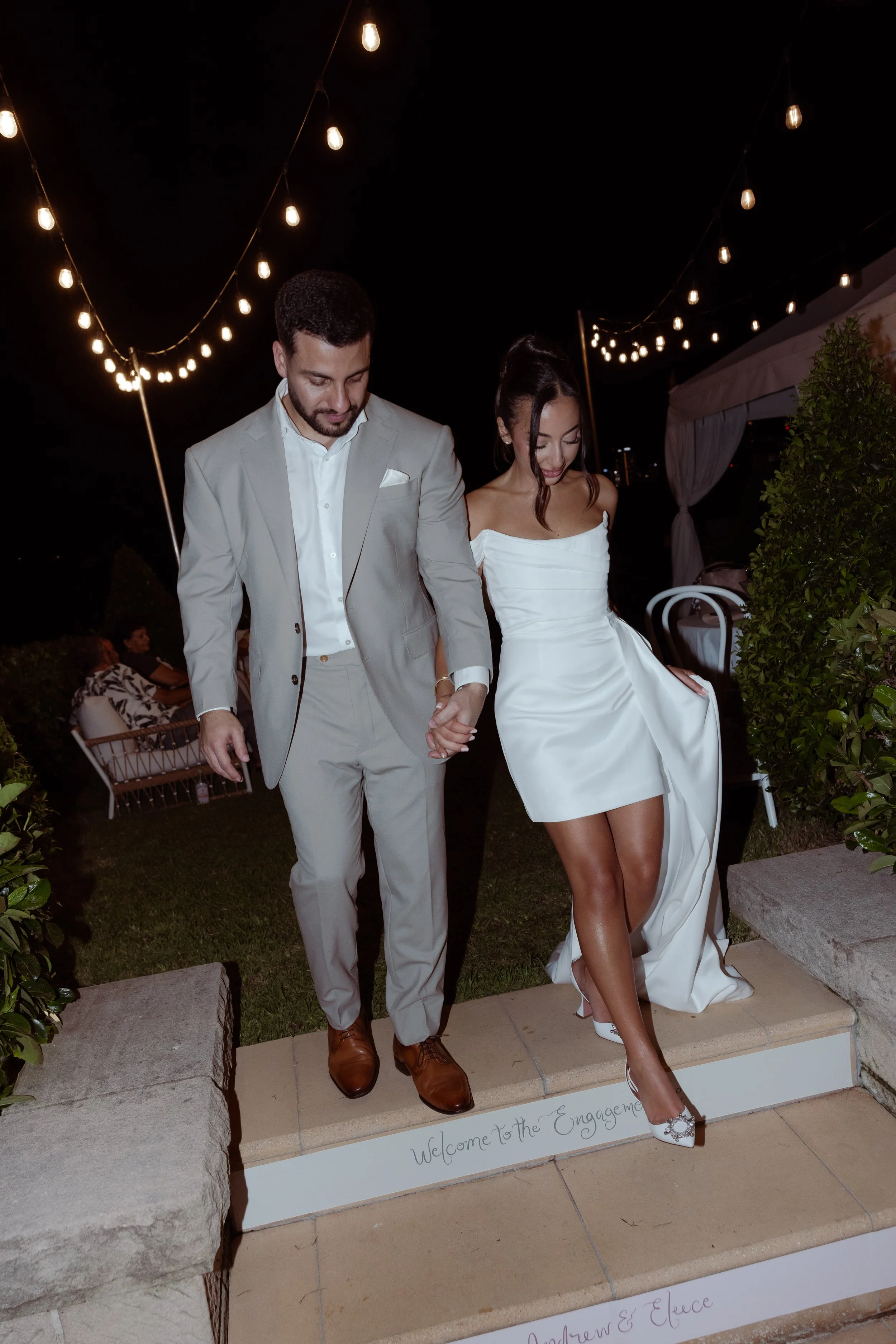 A couple dressed in wedding attire is holding hands and walking down stairs at night, with string lights hanging above. The groom is in a light gray suit and the bride in a white dress. There is a sign that reads 'Welcome to the Engagement' at the bottom of the stairs.
