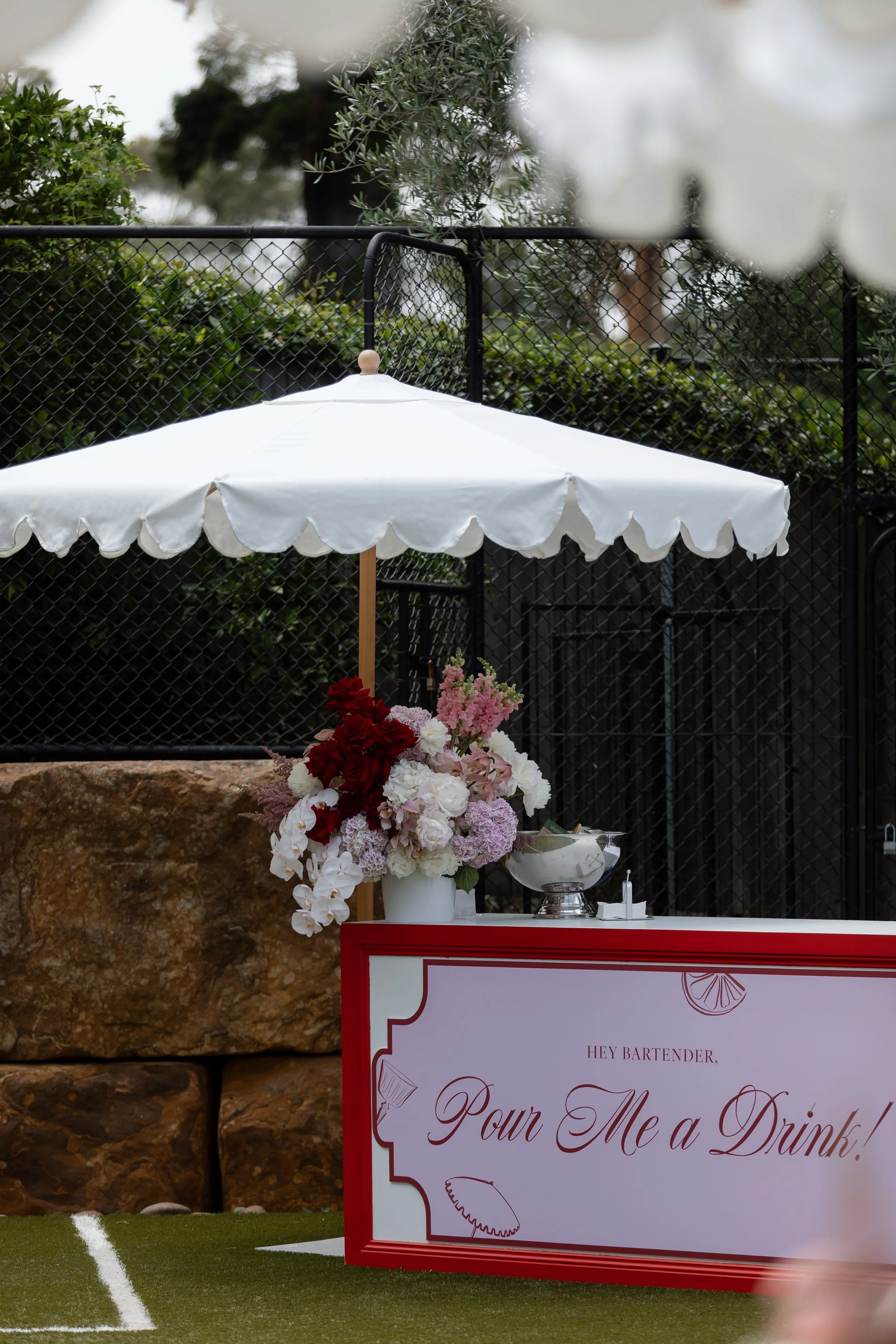 A floral arrangement with pink, white, and red flowers, a white serving dish, and a welcome sign that says 'Pour Me a Drink' on a red-bordered board, placed on a green lawn with a white line, background includes a black fence, trees, and rocks.
