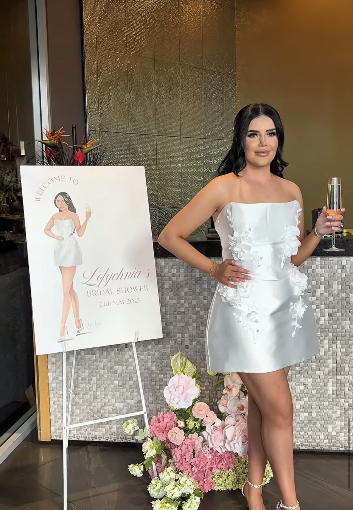 A woman in a white bridal gown holding a glass of champagne, standing next to a sign with a custom illustration of herself for a bridal shower, decorated with pink and white flowers.