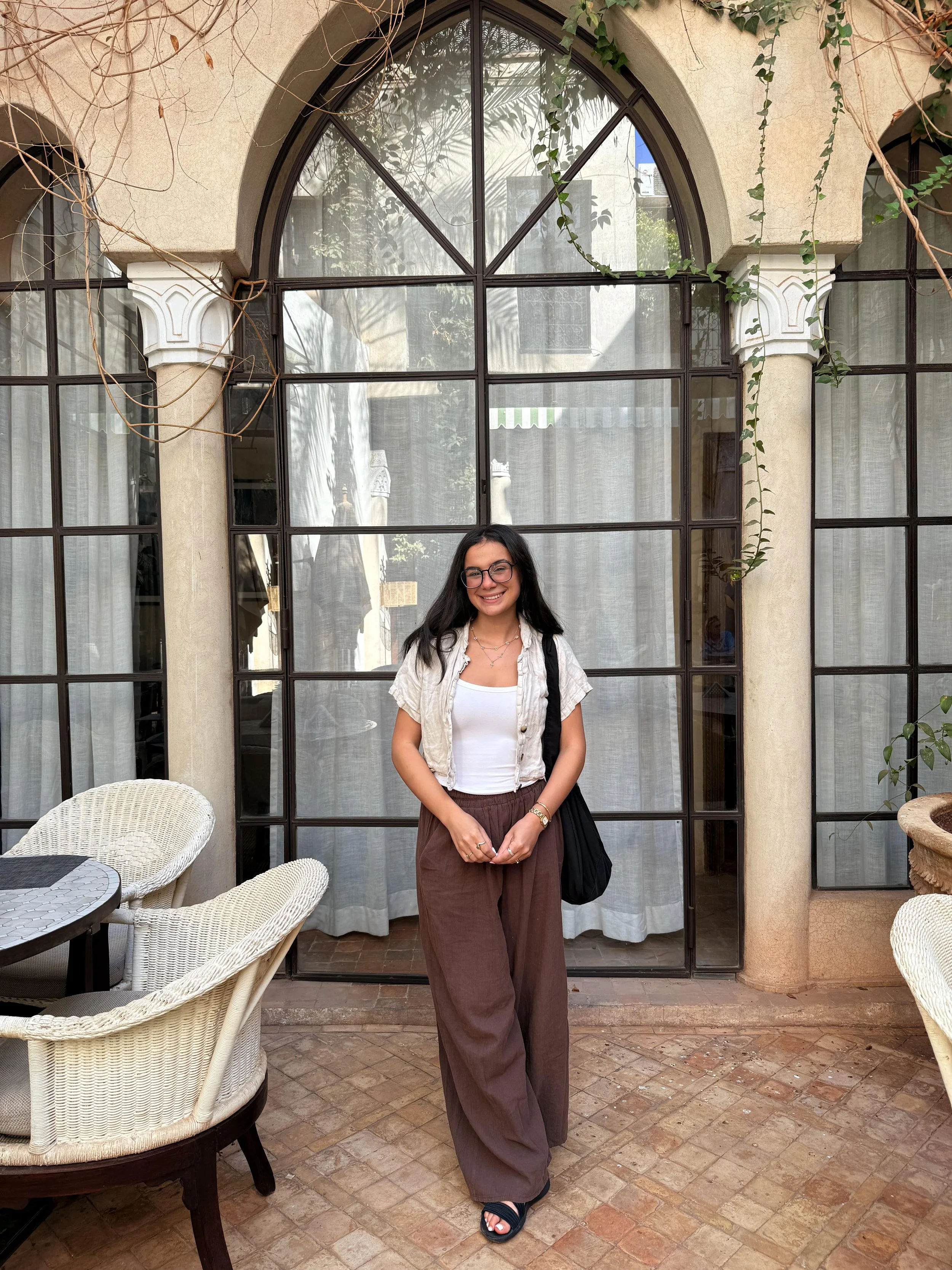 A woman with long dark hair, glasses, and a friendly smile standing in front of a large arched window with curtains. She is wearing a white top, a beige short-sleeve jacket, baggy brown pants, and black sandals, holding a black bag on her left shoulder. The background includes outdoor furniture, potted plants, and a stone patio.