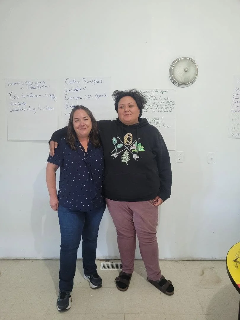 Two women standing close together indoors, smiling, with one arm around each other's shoulders. Whiteboards with handwritten notes are visible on the wall behind them, and a wall-mounted clock is above them.