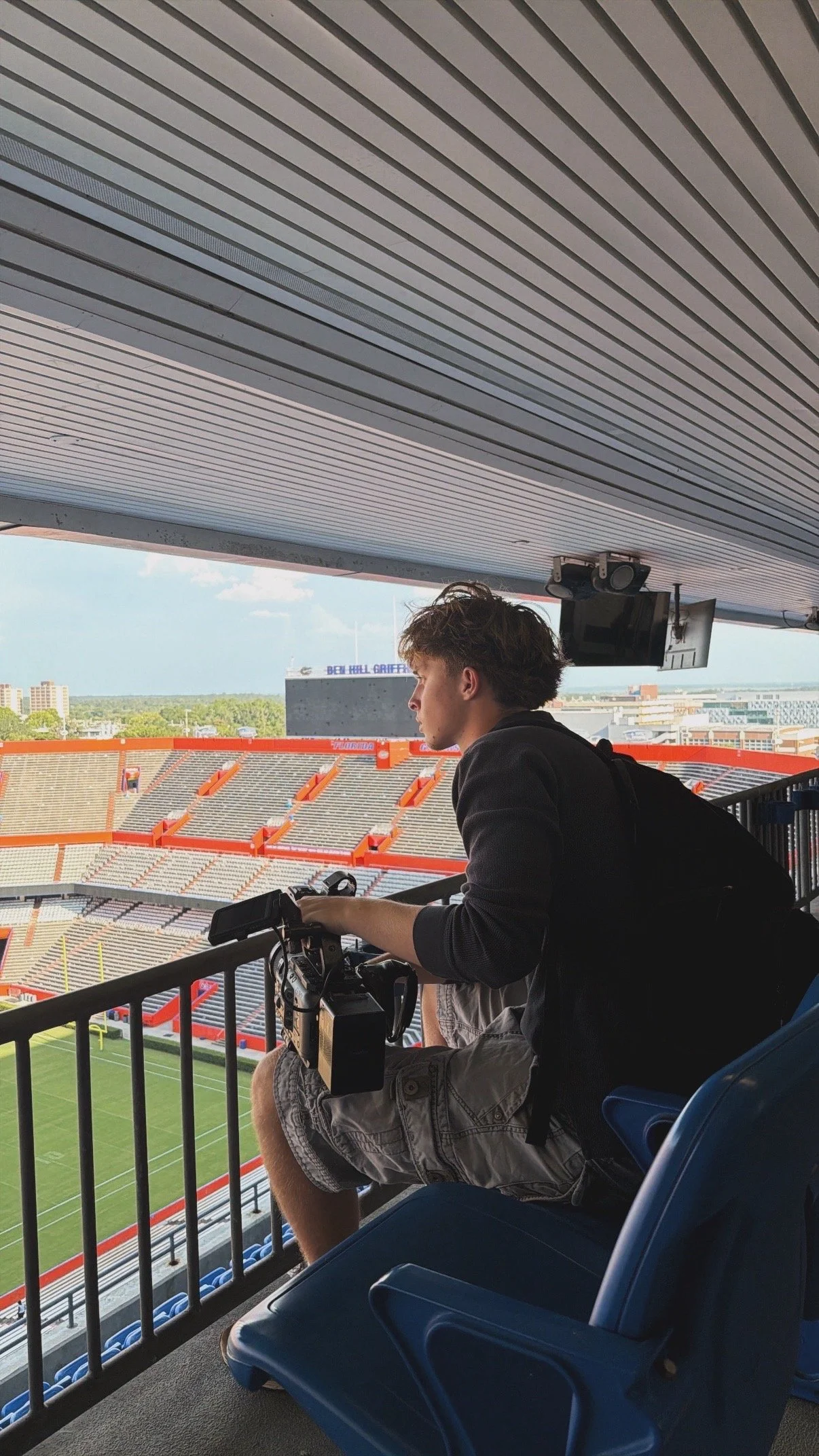 Young man sitting in a stadium watching a football game, with a camera and equipment in front of him, and a view of the field and empty bleachers in the background.