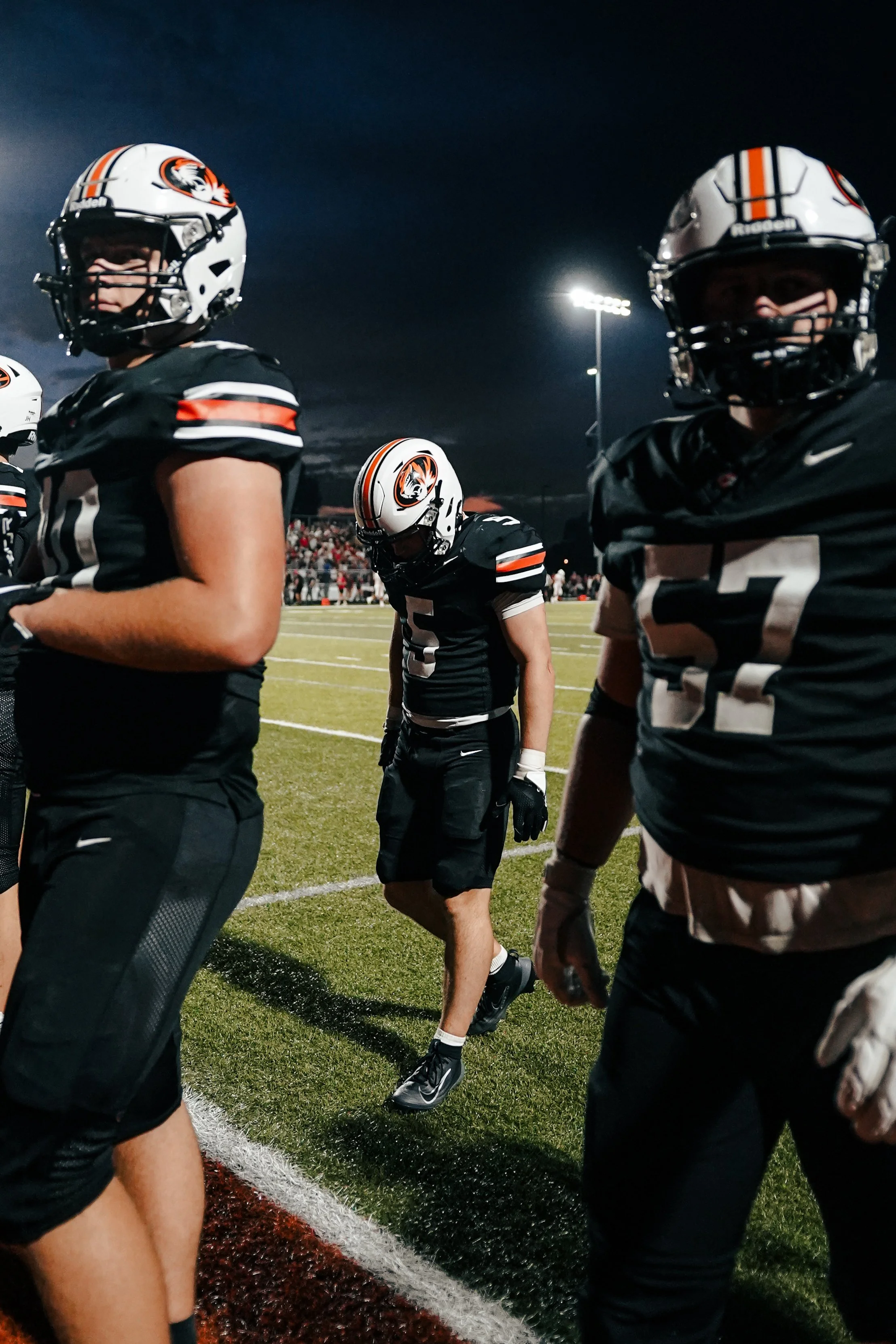 A group of football players wearing helmets and black uniforms with orange and white accents, standing on a football field at night.