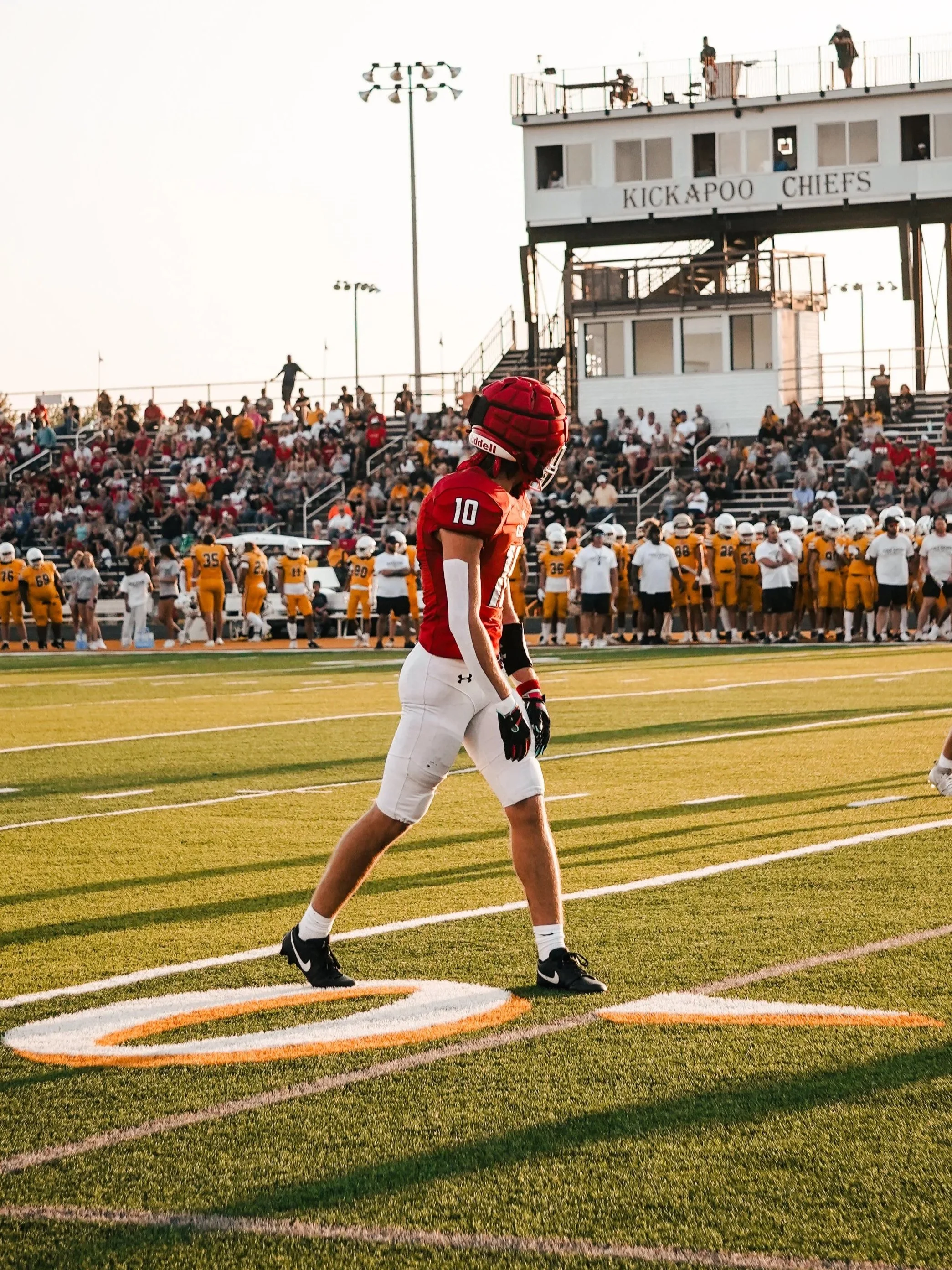 A football player in a red uniform with the number 10 walking along the sideline of a football field during a game, with spectators in the stands and a press box above.