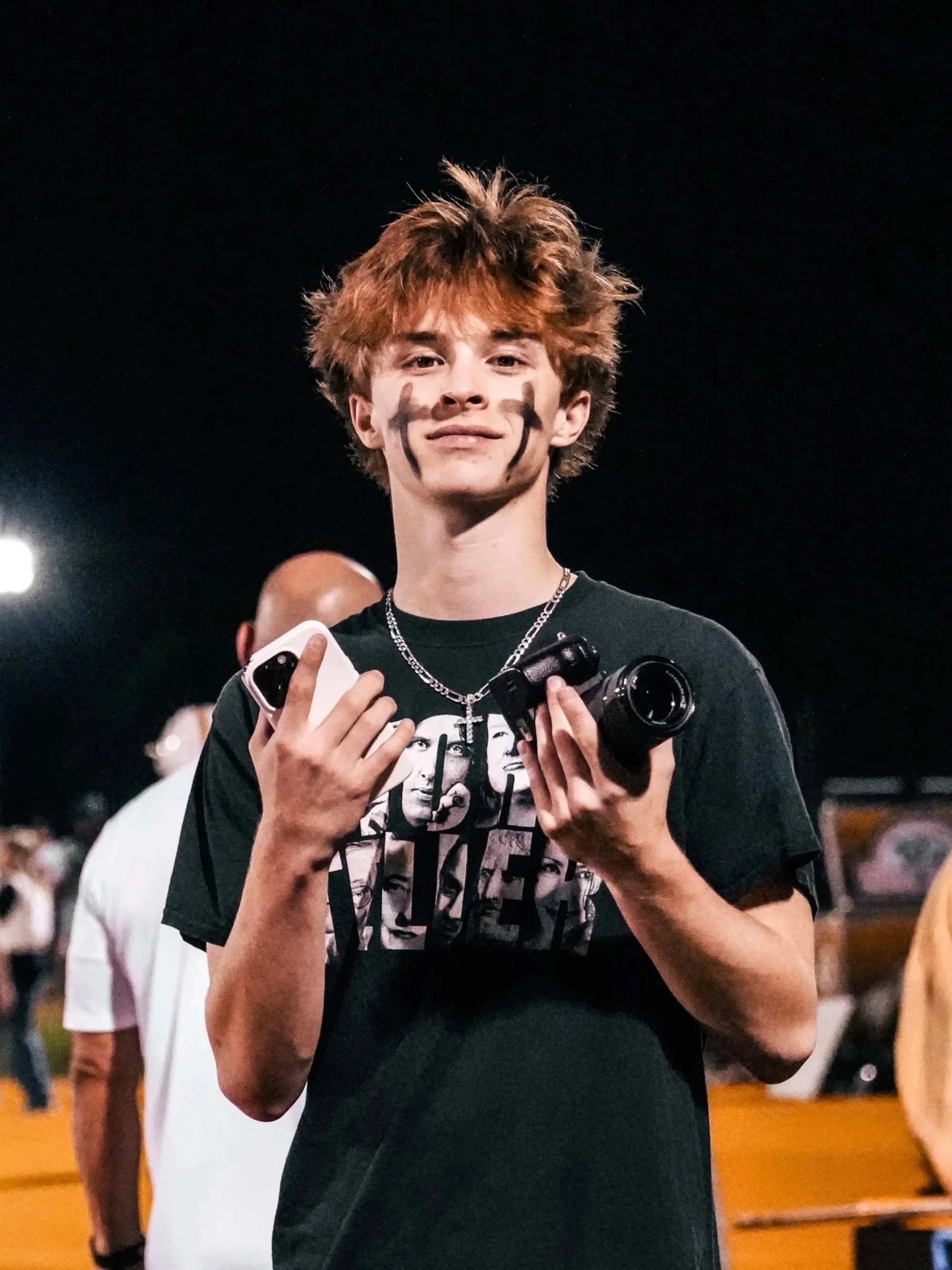 Young man with messy red hair, black paint or marker on his face, holding a camera and a smartphone, standing in an indoor location with people in the background.