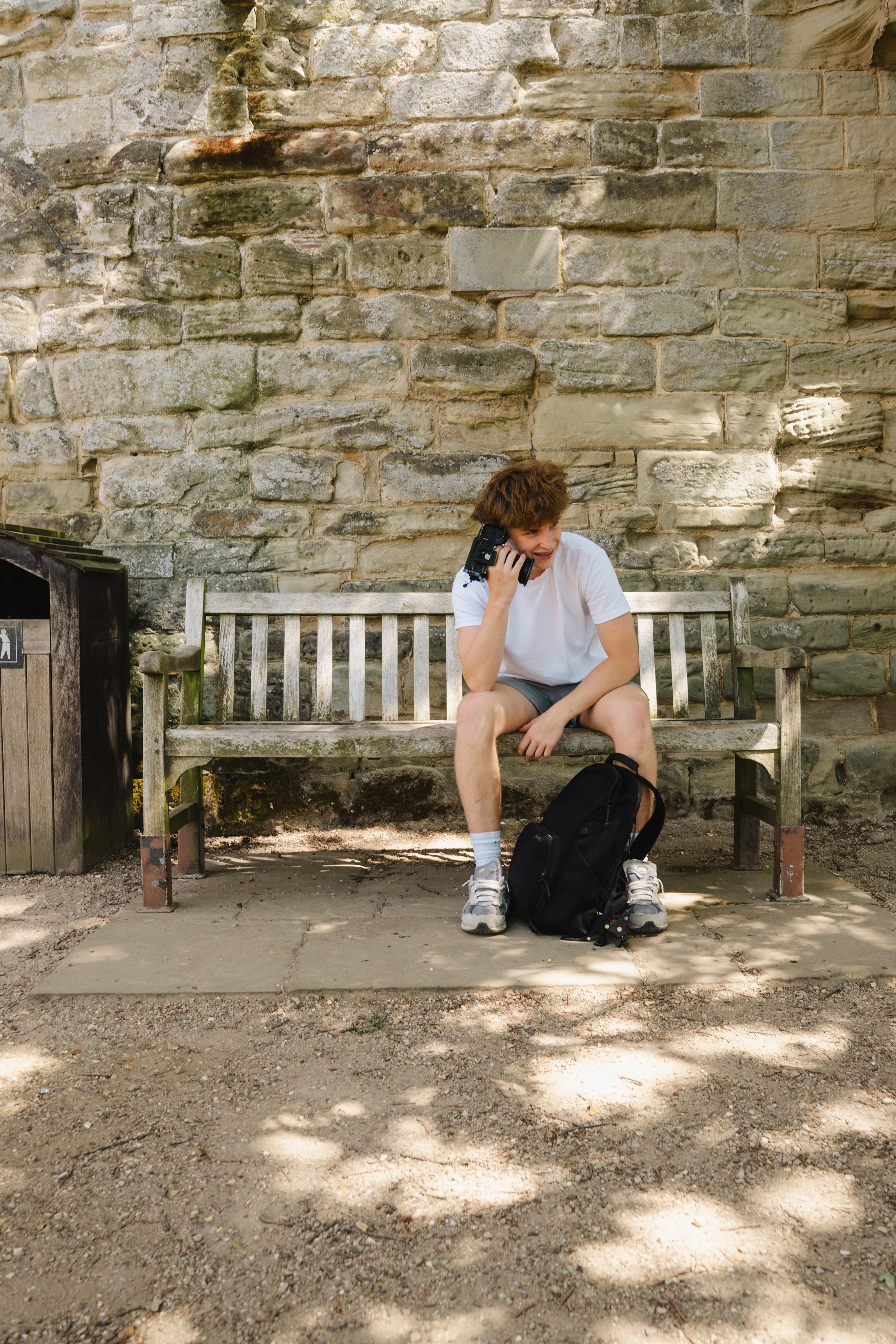 A young man with curly brown hair wearing a white T-shirt, gray shorts, white sneakers, and gray socks, sitting on a weathered wooden bench in front of a stone wall. He has a black backpack on the ground in front of him and is holding a camera to his ear, smiling. Sunlight filters through trees, casting shadows on the ground.