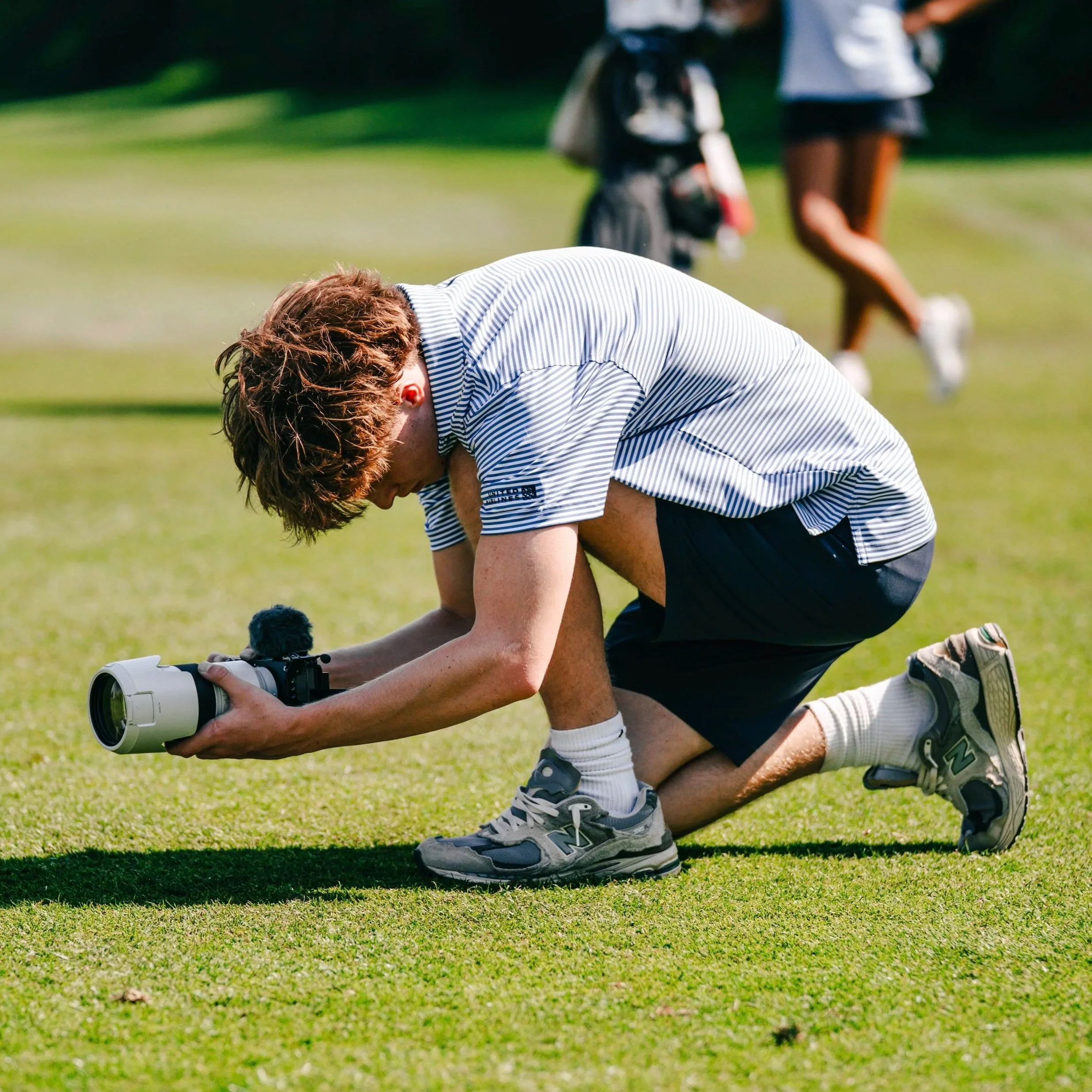 A person kneeling on a golf course, setting up a golf ball with a rangefinder or golf club, while another person stands nearby in the background.