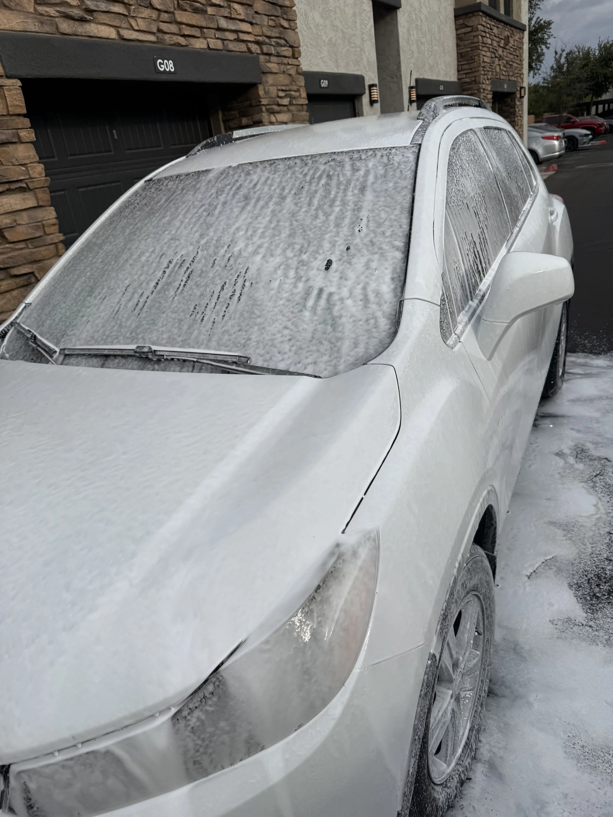 A white car covered in soap foam parked outside a building with a stone and stucco exterior during rainy weather.