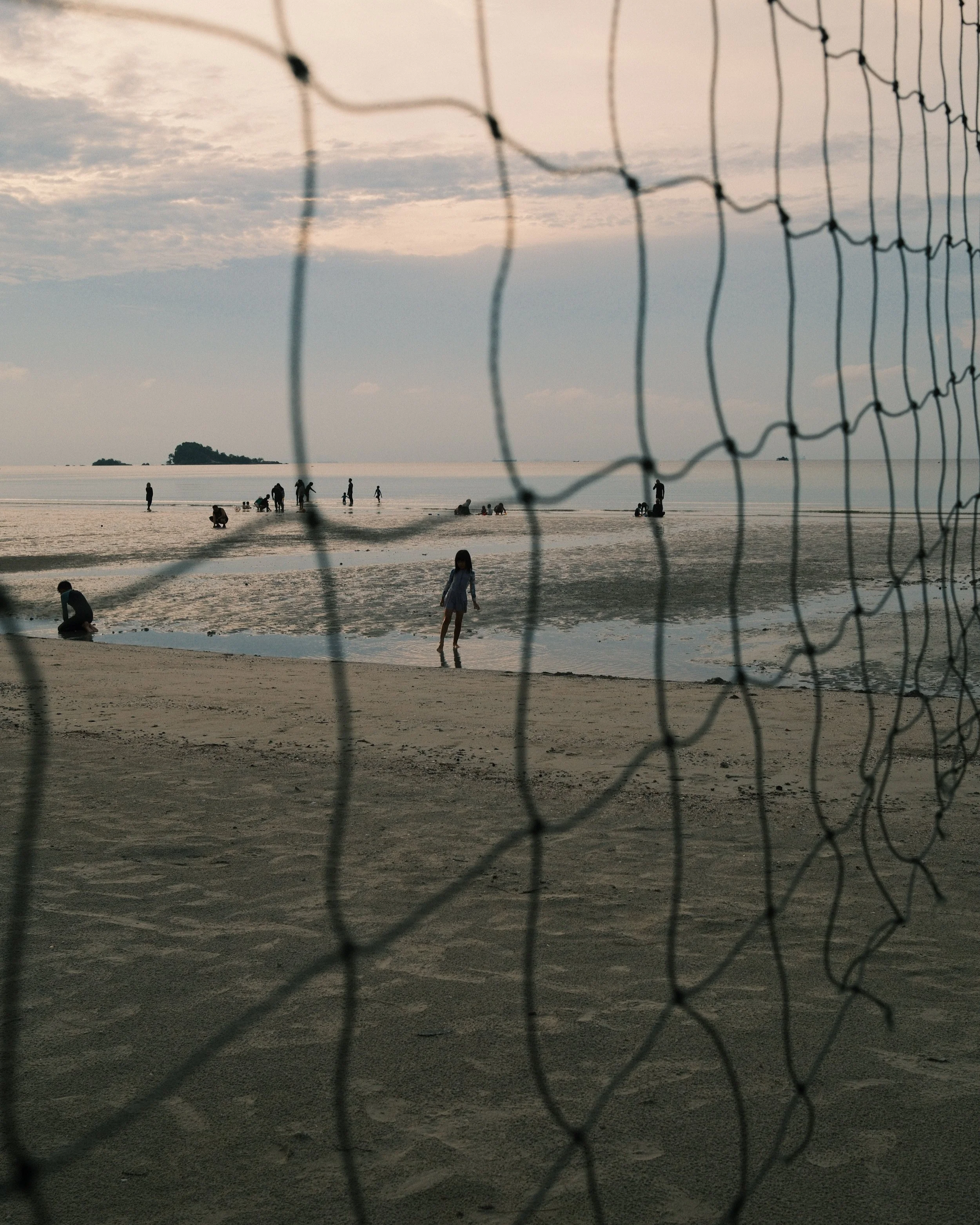 Views of a beach with people playing, wading, and sitting. A wire fence in the foreground partially obscures the scene. An island is visible in the background, under a cloudy sky at dusk.