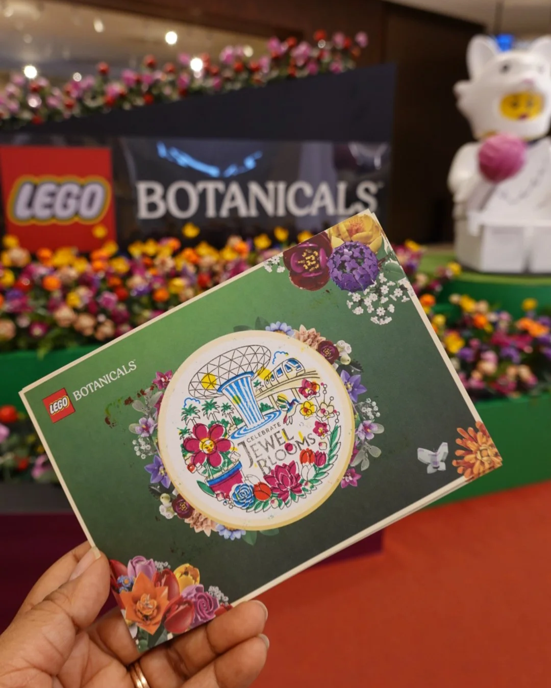 A person holds a green brochure with colorful flowers and the words 'Celebrate Jewel of Blooms' in front of a display of various small flowers at a LEGO botanicals exhibit.