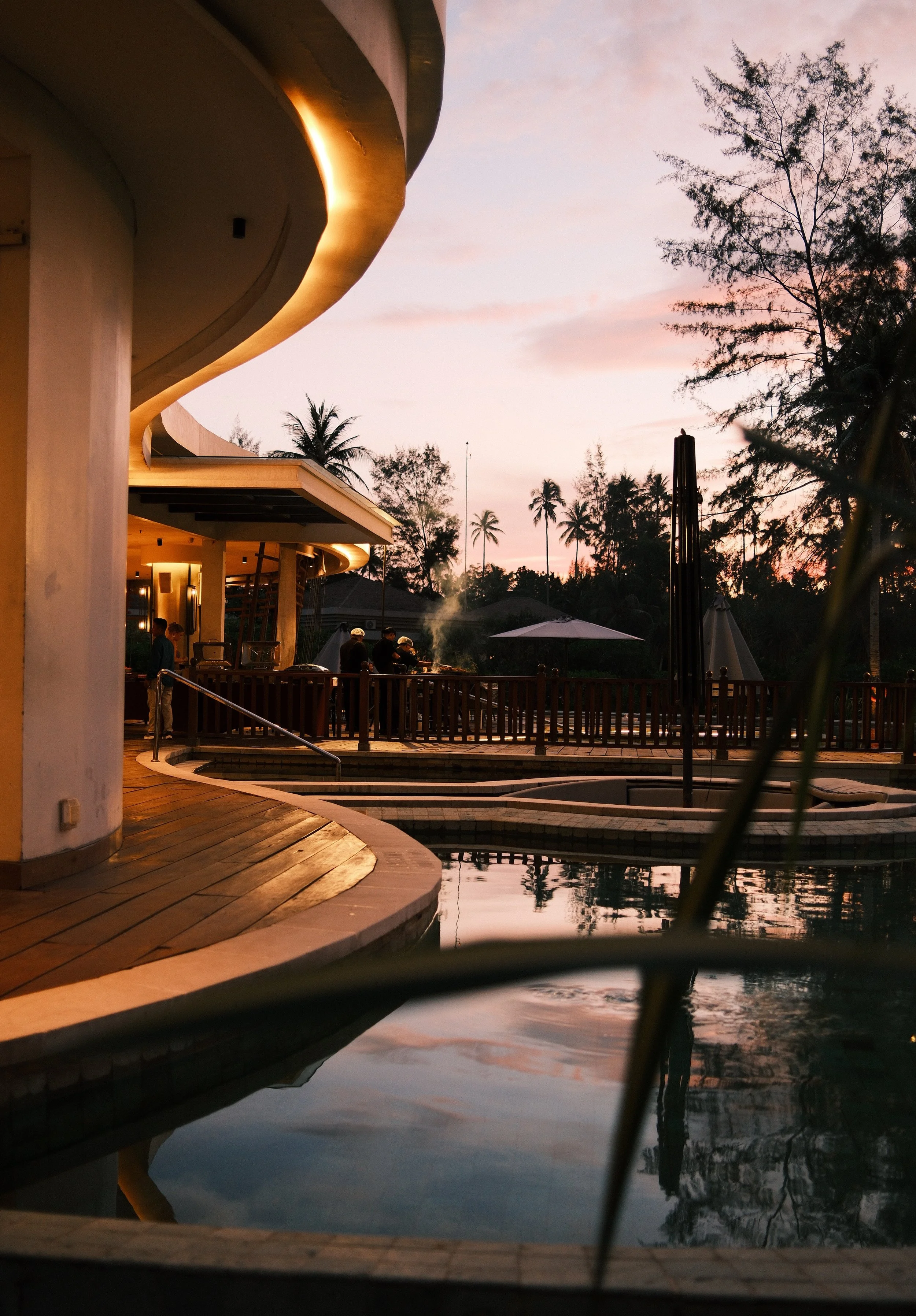 A poolside scene at sunset with a modern building, a wooden deck, people grilling, and palm trees in the background under a pinkish sky.